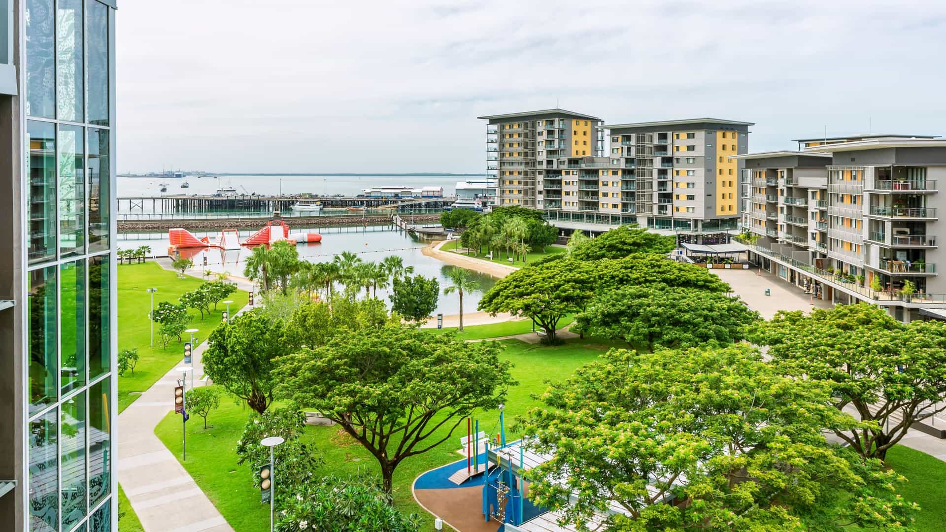 A bright daytime photo of the Darwin Waterfront in Australia, showcasing the man made saltwater recreation lagoon and surrounding modern high rise apartment buildings and palms under a clear blue sky.