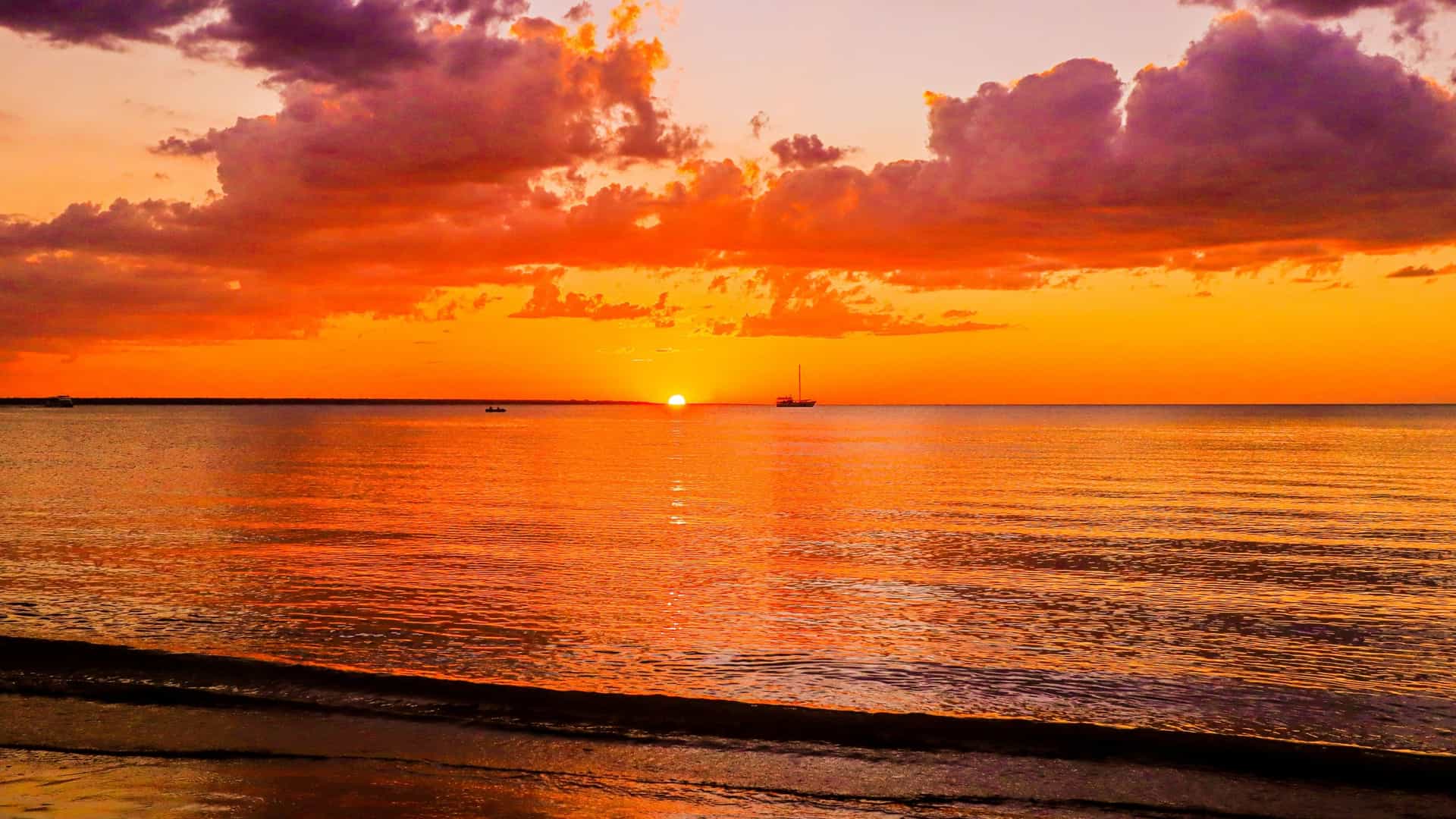 A wide view of a vibrant sunset over Mindil Beach in Darwin, Australia. The sky is ablaze with warm colors of orange, red, and yellow, reflecting brightly on the wet, expansive sand and shallow water. Silhouettes of people are scattered across the beach enjoying the view