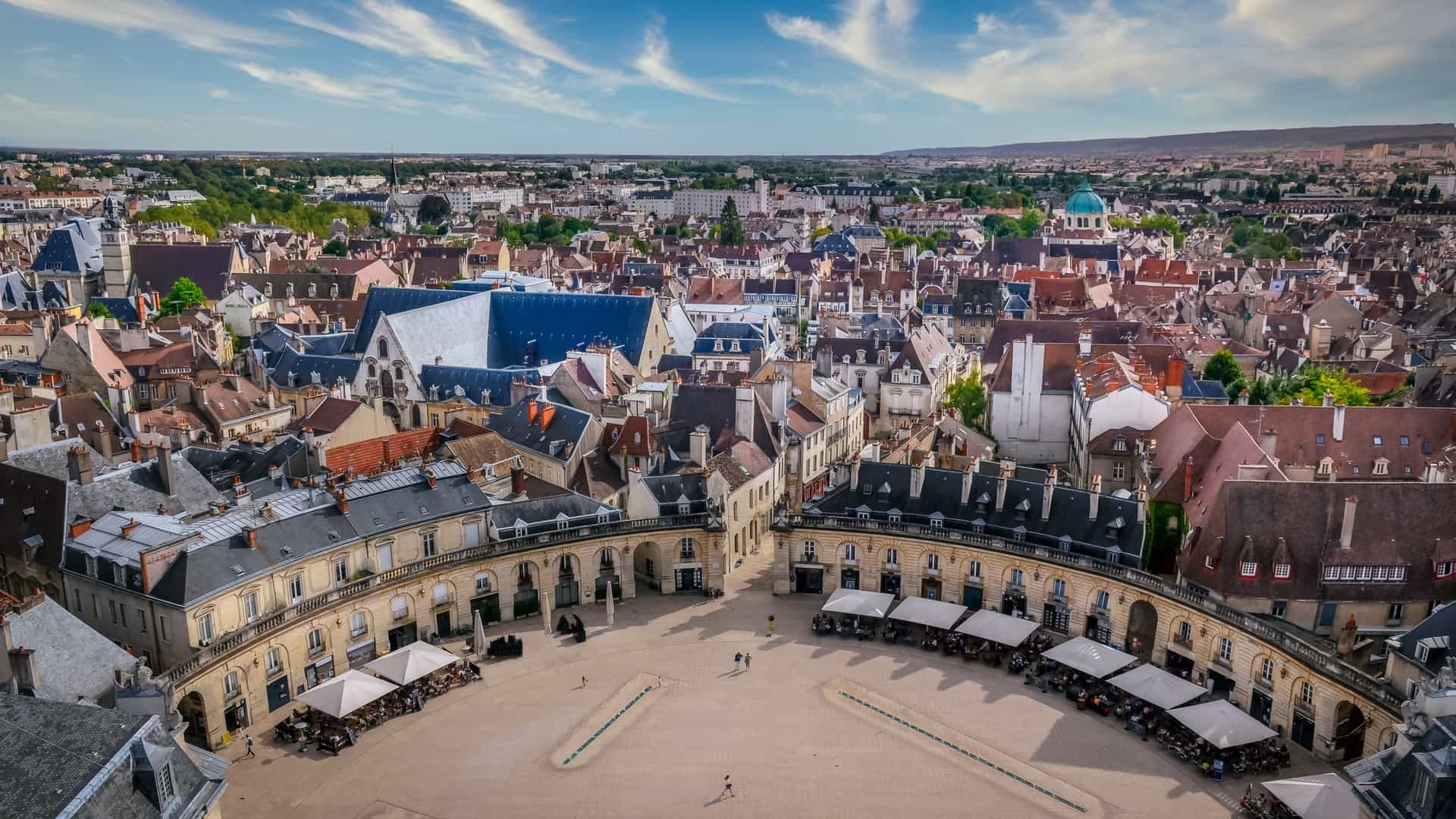 A cityscape view of Place de la Libération in Dijon, France. The semicircular, pedestrian square is surrounded by grand, classical buildings, including the Palace of the Dukes of Burgundy, under a clear blue sky.