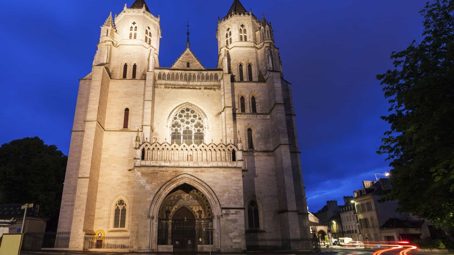 A close up view of the facade and bell tower of the Gothic style St. Bénigne Cathedral in Dijon, France. The ornate stonework is visible, along with the tall, pointed spire that rises above the rooftops.
