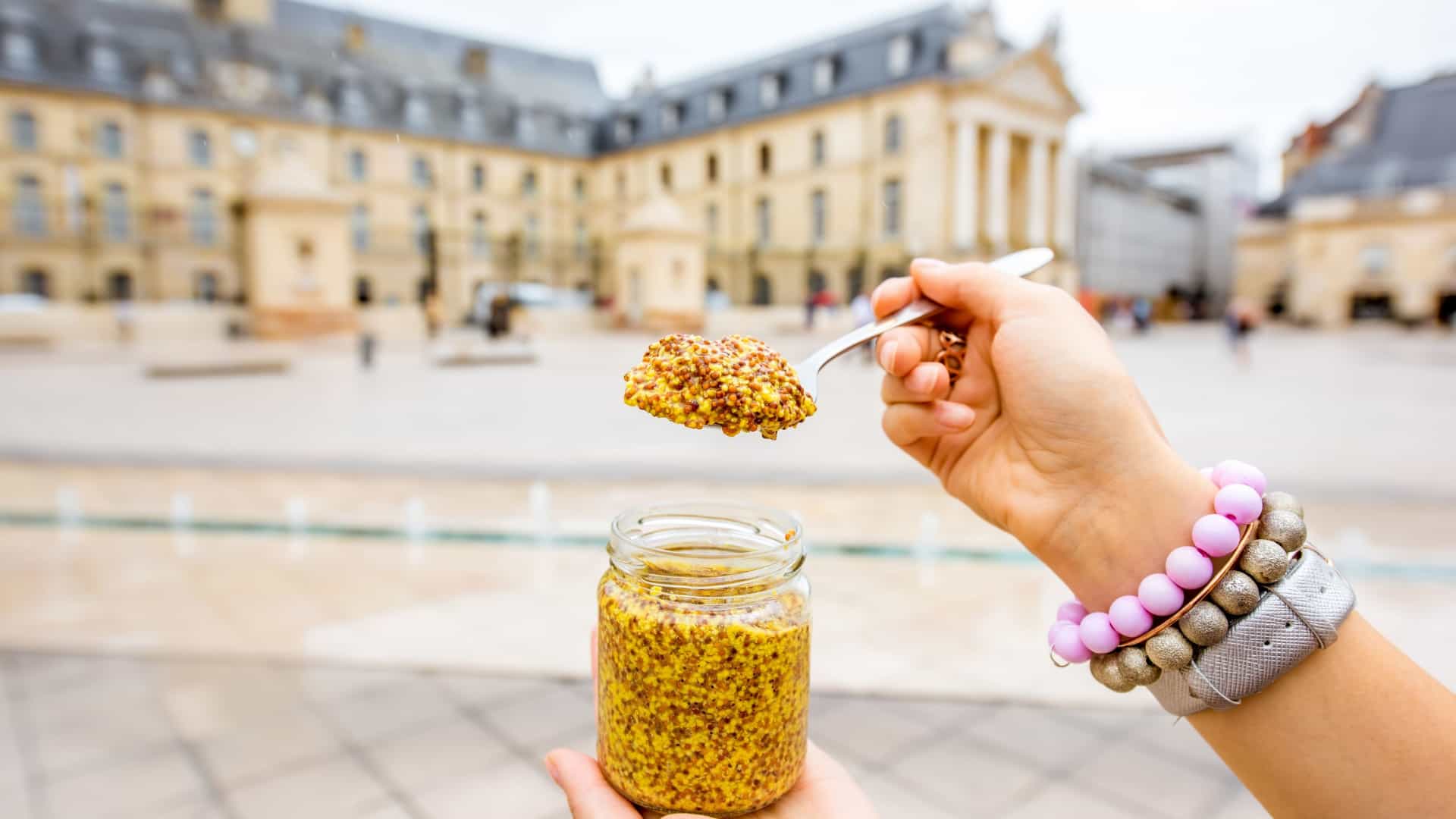 A close up shot of a hand holding a small glass jar of authentic, famous Dijon mustard, a popular French gourmet culinary condiment used in cooking.