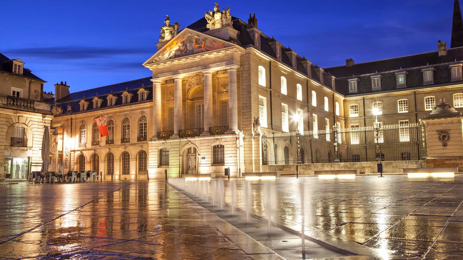 A captivating dusk photograph of the semicircular Liberation Square and the magnificent Palace of the Dukes of Burgundy in Dijon, France, with the building beautifully illuminated against the blue sky and reflections on the central fountain.
