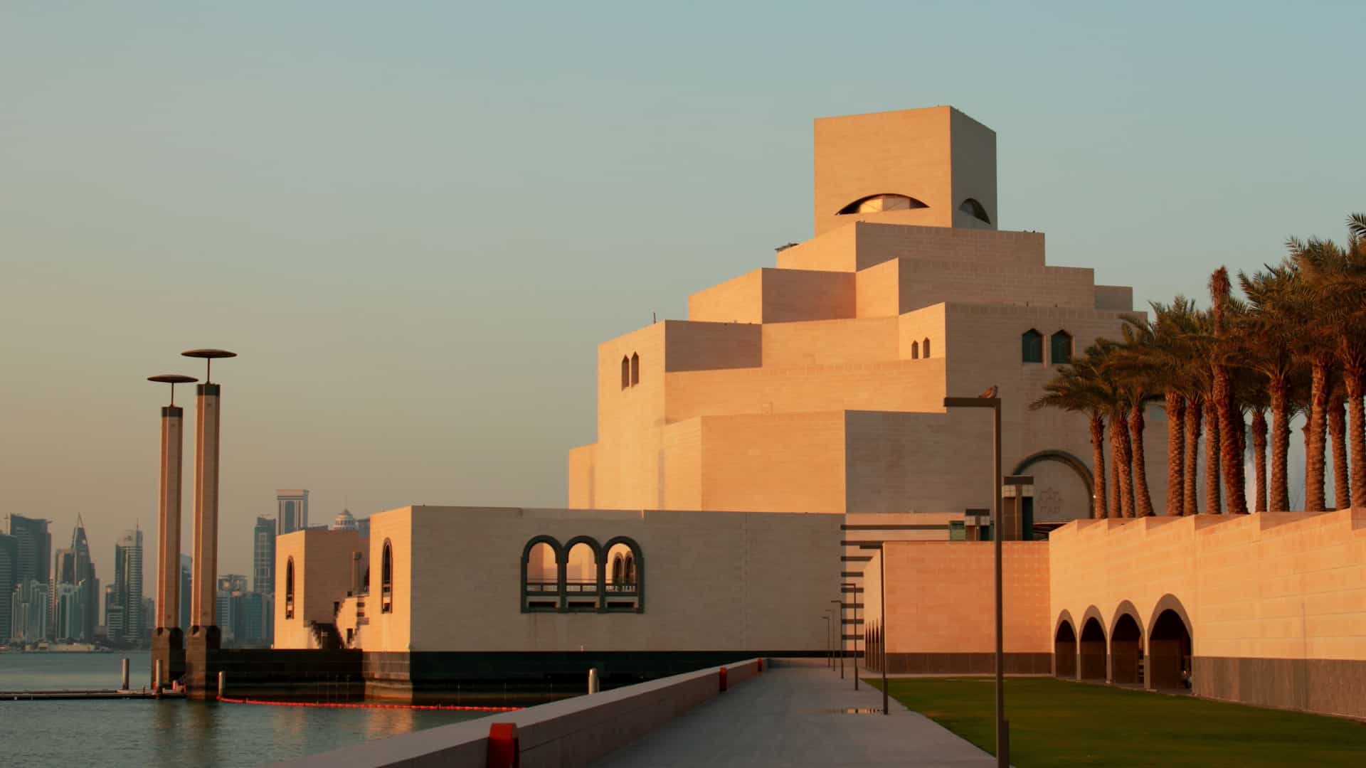 The Museum of Islamic Art in Doha, Qatar. The building is a modern architectural masterpiece with a stark, geometric design of stacked cream colored stone blocks, surrounded by water and set against a clear blue sky.