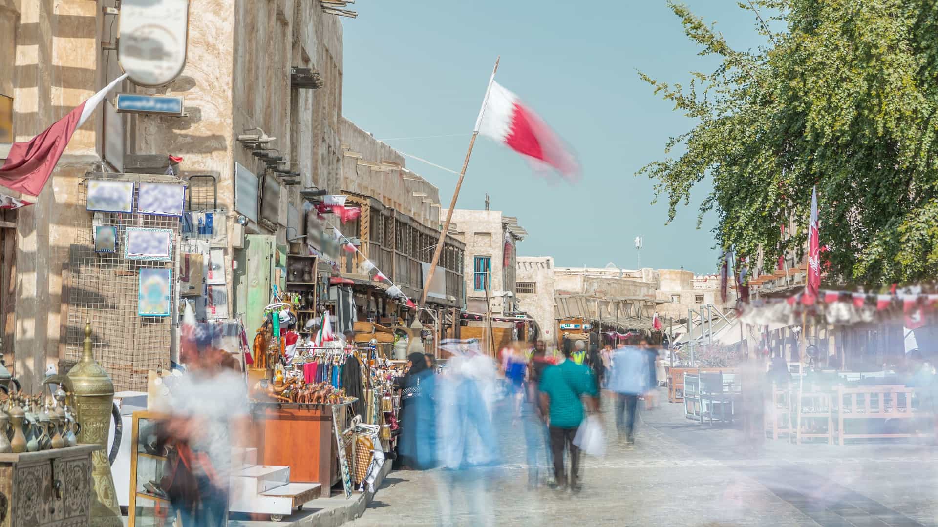 A time lapse of Souq Waqif, a popular marketplace in Doha, Qatar, transitioning from day to night. The image shows the traditional architecture of the souq and the brightly lit buildings and shops at night, with an illuminated Islamic cultural center visible in the background.