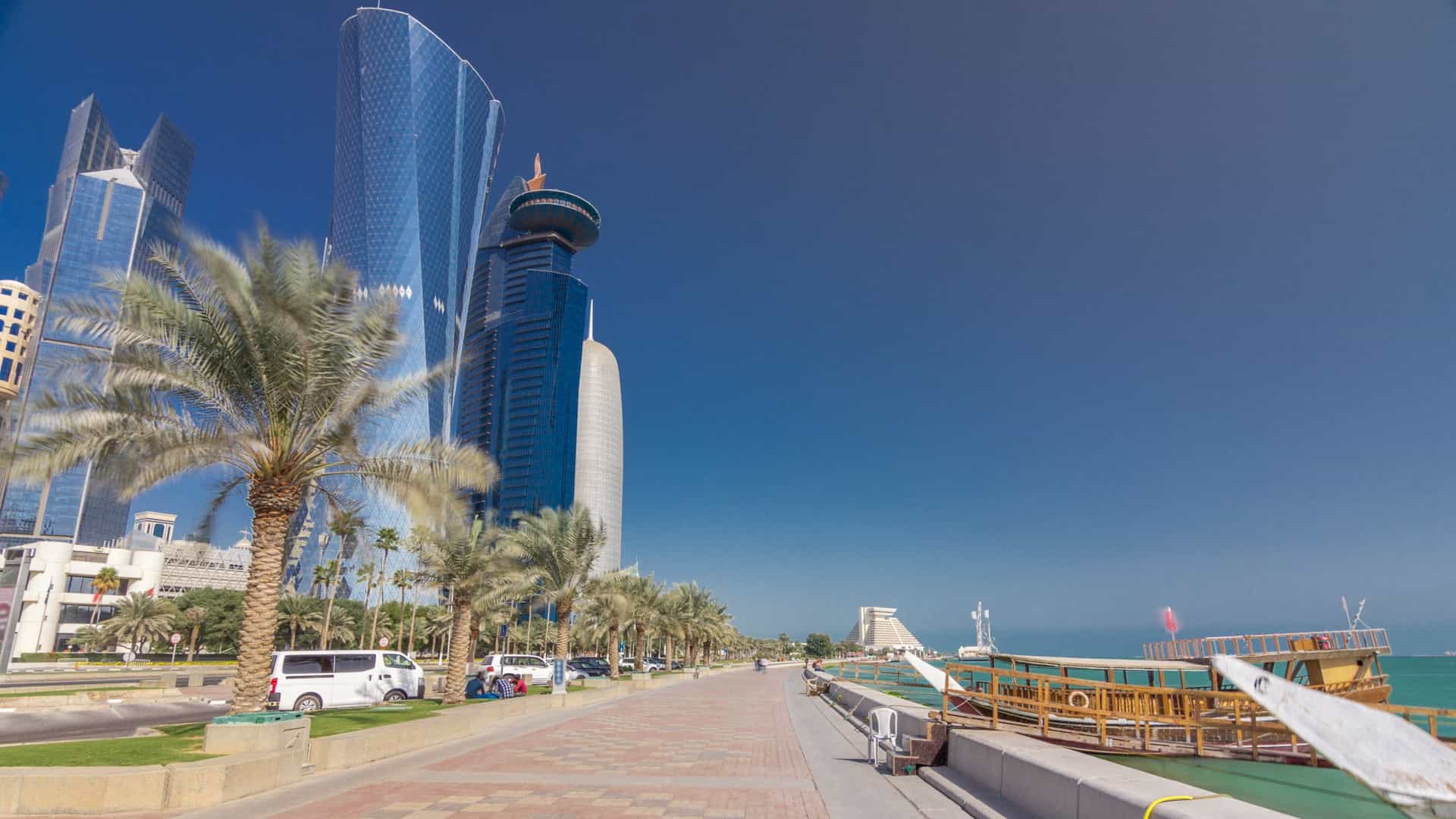 A view of the Doha Corniche waterfront promenade, with the modern skyline of the West Bay district across the bay. The image features a long, curving walkway along the water, with the distinct, illuminated skyscrapers of Doha's business district in the background.