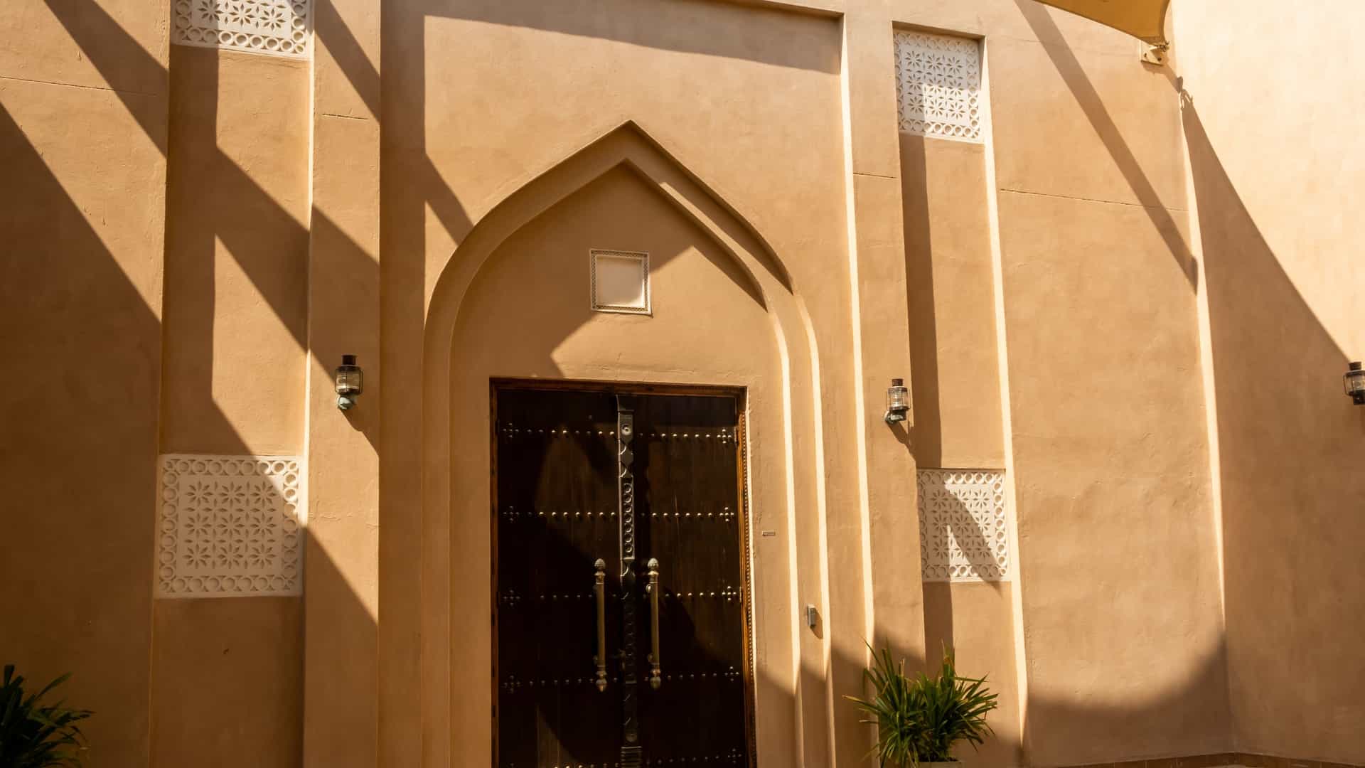 A view of a building at Katara Cultural Village in Doha, Qatar. The architecture is inspired by traditional Qatari design, featuring sand colored, low rise structures with intricate patterns and details, set against a backdrop of water and a clear blue sky.
