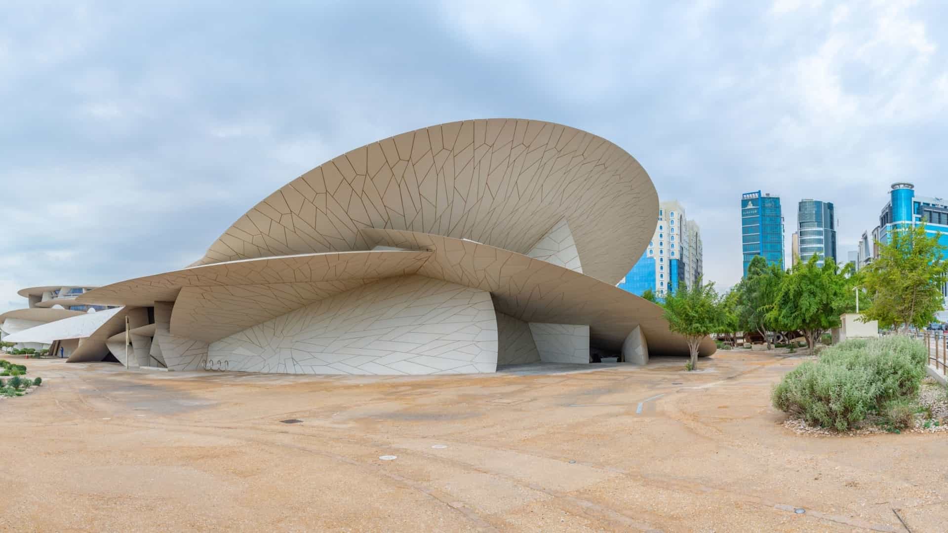 A wide exterior view of the National Museum of Qatar in Doha, designed by Jean Nouvel. The building's striking, modern architecture is inspired by the crystalline structure of a desert rose, featuring numerous large, interlocking, disc shaped elements in a sandy beige color against a blue sky.