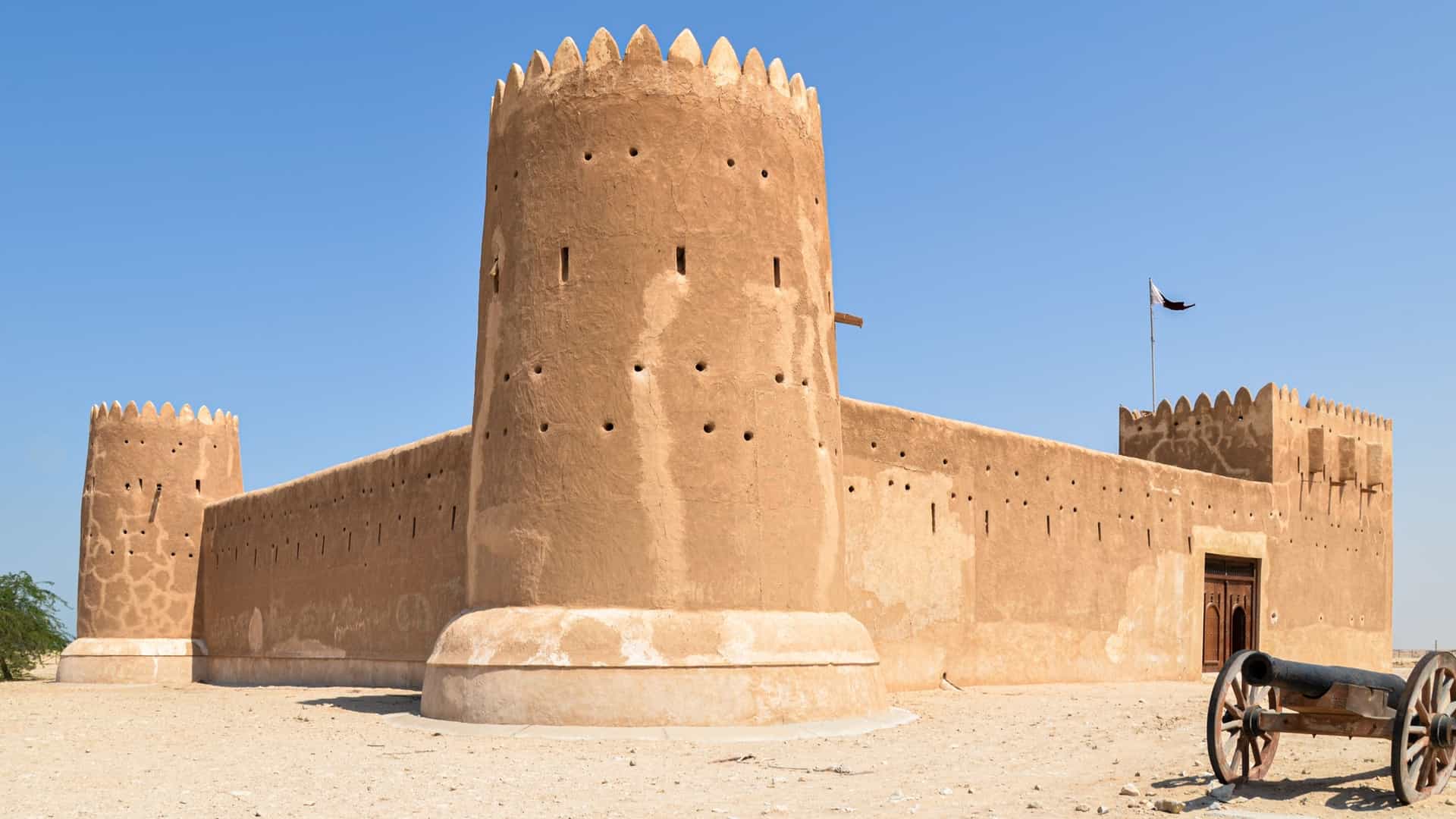 An external view of the historical Al Zubarah Fort in Qatar. The fort, a traditional Qatari military fortress, is a square structure with thick, beige colored walls and four corner towers. It stands in a desert landscape under a clear blue sky. The fort is a key part of the Al Zubarah Archaeological Site, a UNESCO World Heritage site.