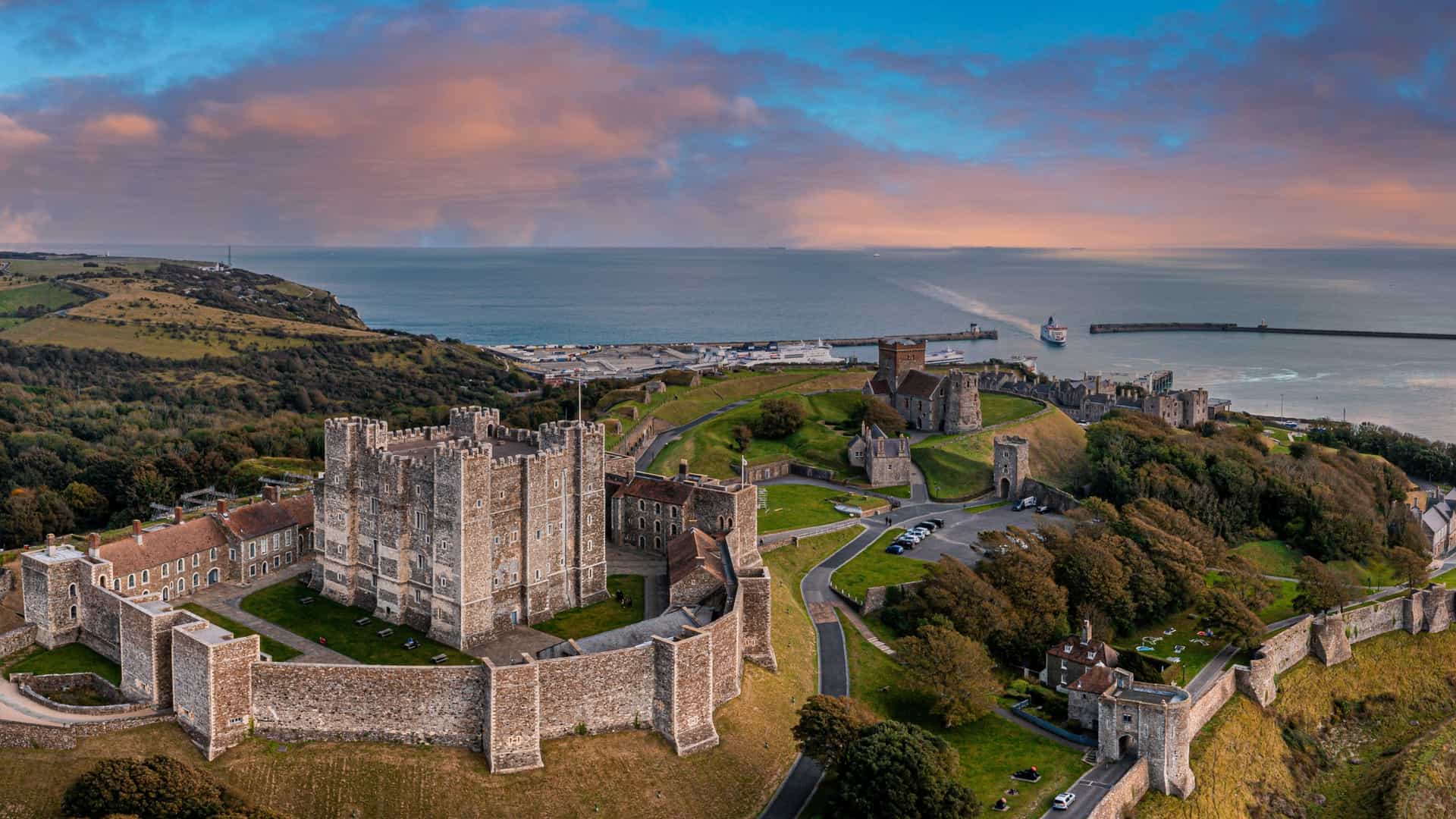 An aerial view of the historic Dover Castle in Kent, England. The medieval fortress, with its prominent keep and surrounding walls, is located on a hill overlooking the town of Dover and the English Channel. The image shows the castle complex on a green landscape, with the coastline and ocean visible in the background.