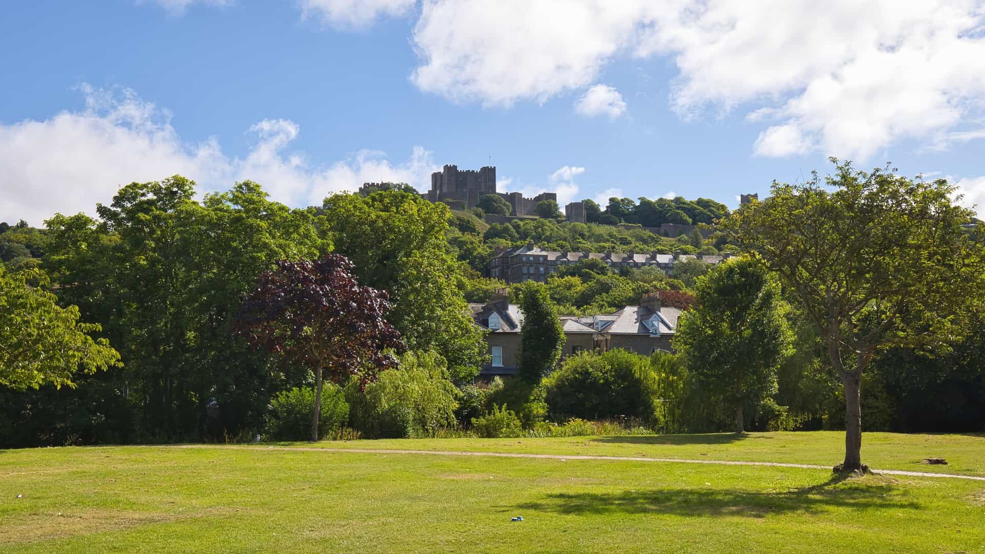 An aerial, sunny view of Pencester Gardens, a green urban park in Dover, England. The image shows wide, manicured lawns, a winding path, and a circular bandstand pavilion, surrounded by mature trees and the dense urban environment.