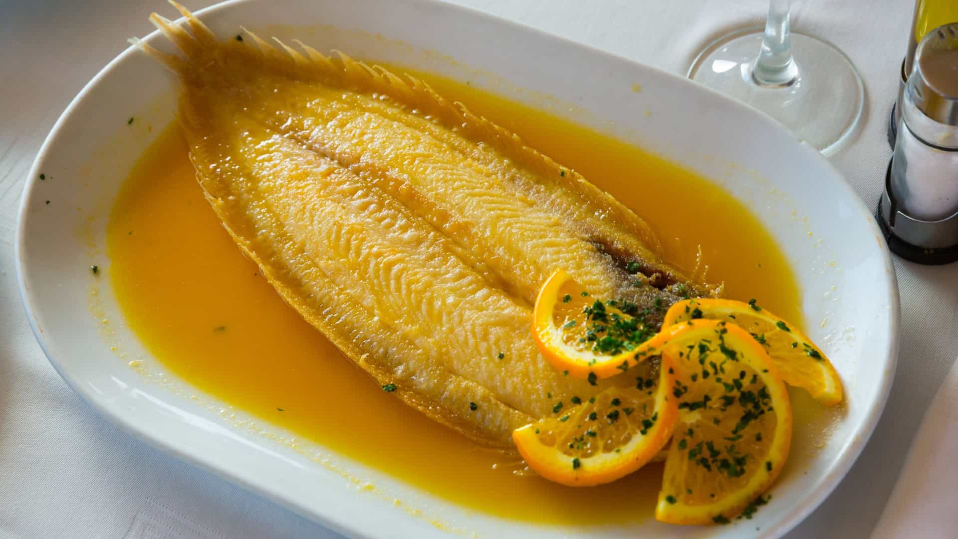 A fillet of light colored, cooked Dover sole fish, drizzled with a rich, bright orange sauce and garnished with capers or herbs, served on a round white dining plate, suggesting an elegant seafood dish.