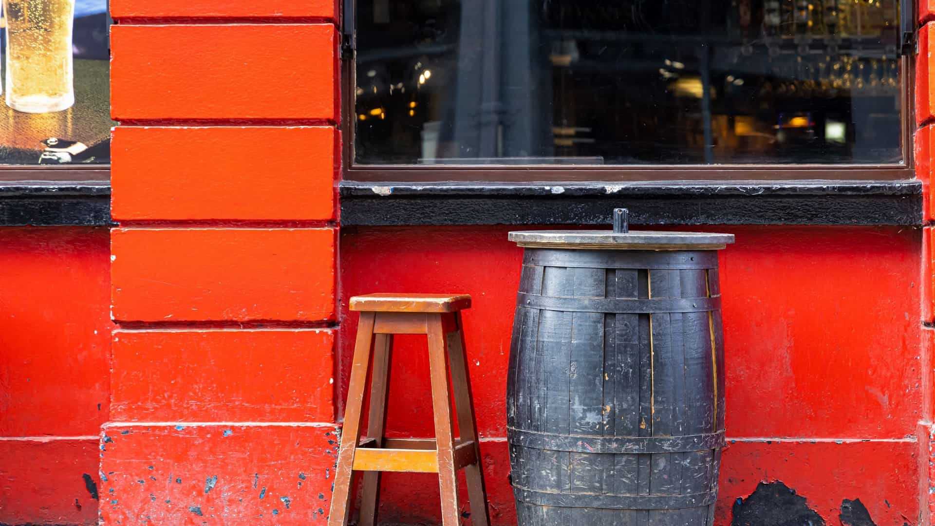 A close up of a traditional outdoor seating area in Dublin's Temple Bar district. The image features a dark, wooden beer barrel (used as a table) and a small wooden stool set against a vibrant, fire engine red wall and a black trimmed window.