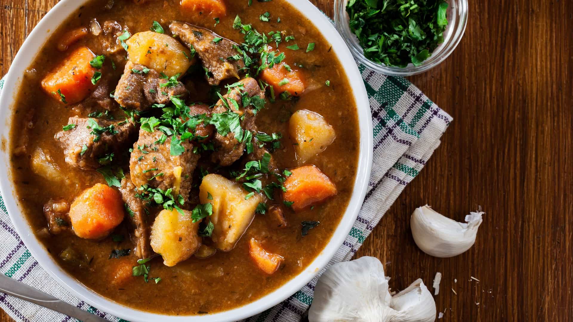 A top down view of a bowl of Irish stew. The hearty, traditional dish contains large, rustic chunks of beef, potatoes, and carrots in a rich, dark gravy. The stew is garnished with fresh, chopped herbs, likely parsley, and is served in a dark colored bowl on a wooden surface.