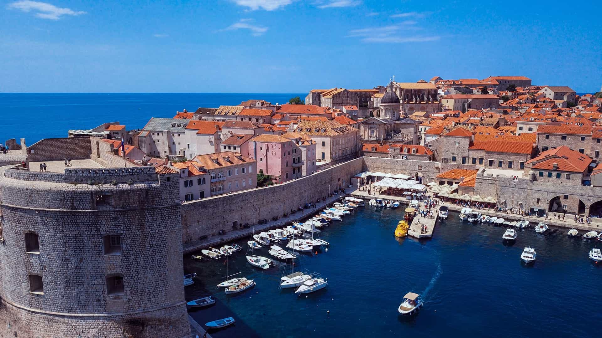 A high angle aerial view of the historic Old Town of Dubrovnik, Croatia, showing a dense cluster of buildings with uniform terracotta red rooftops, encircled entirely by thick, medieval stone city walls. The city is situated against the brilliant blue of the Adriatic Sea, with Fort Lovrijenac visible in the upper part of the image.