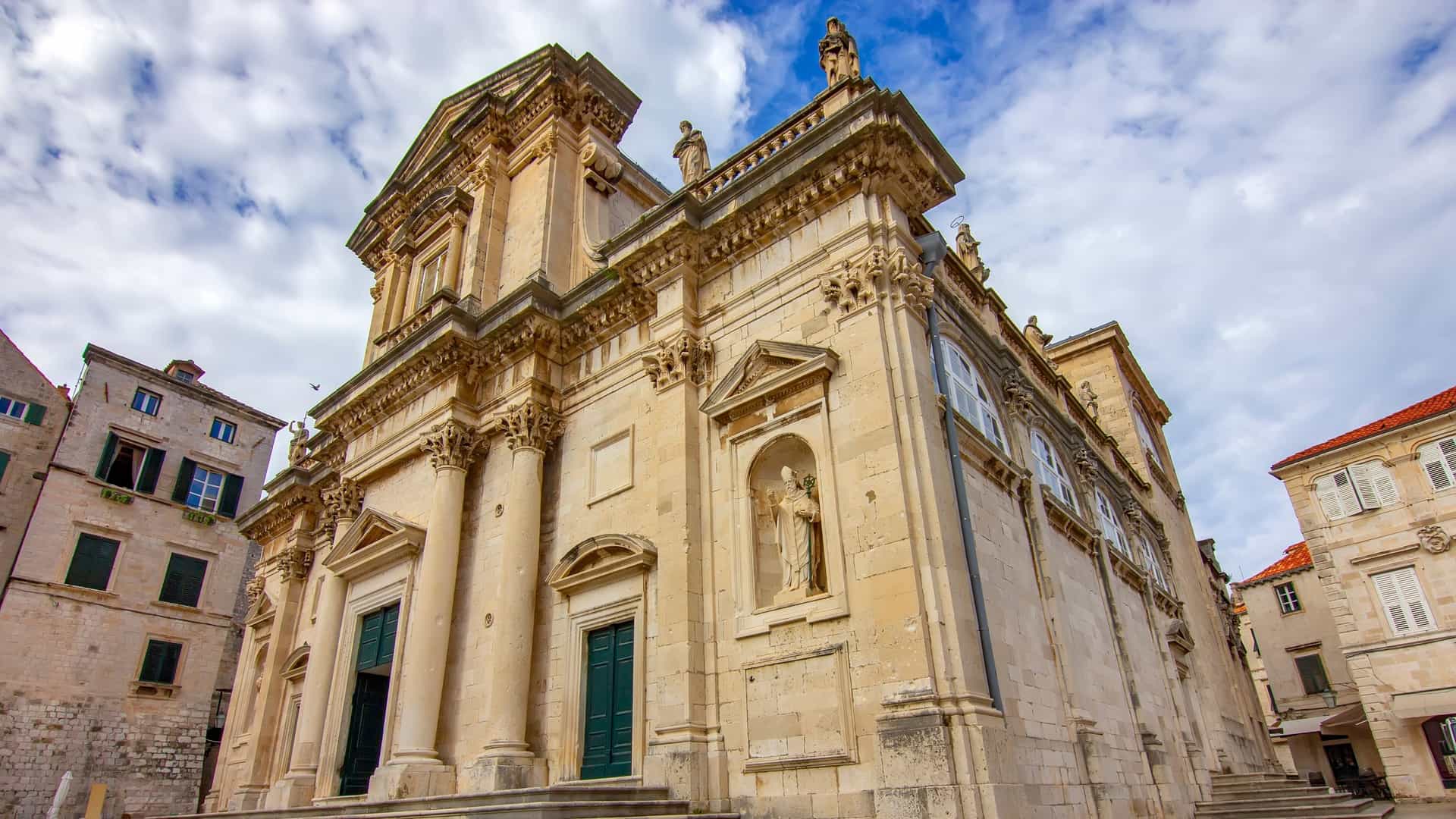 A photograph of the Dubrovnik Cathedral in Croatia. The building is a prominent example of Baroque architecture, featuring a large dome and a stone facade. The image shows the cathedral from an elevated perspective, likely from the city walls, highlighting its position within the historic red roofed Old Town. The sky is bright and clear, and the image captures the intricate details of the dome and the surrounding stone buildings.