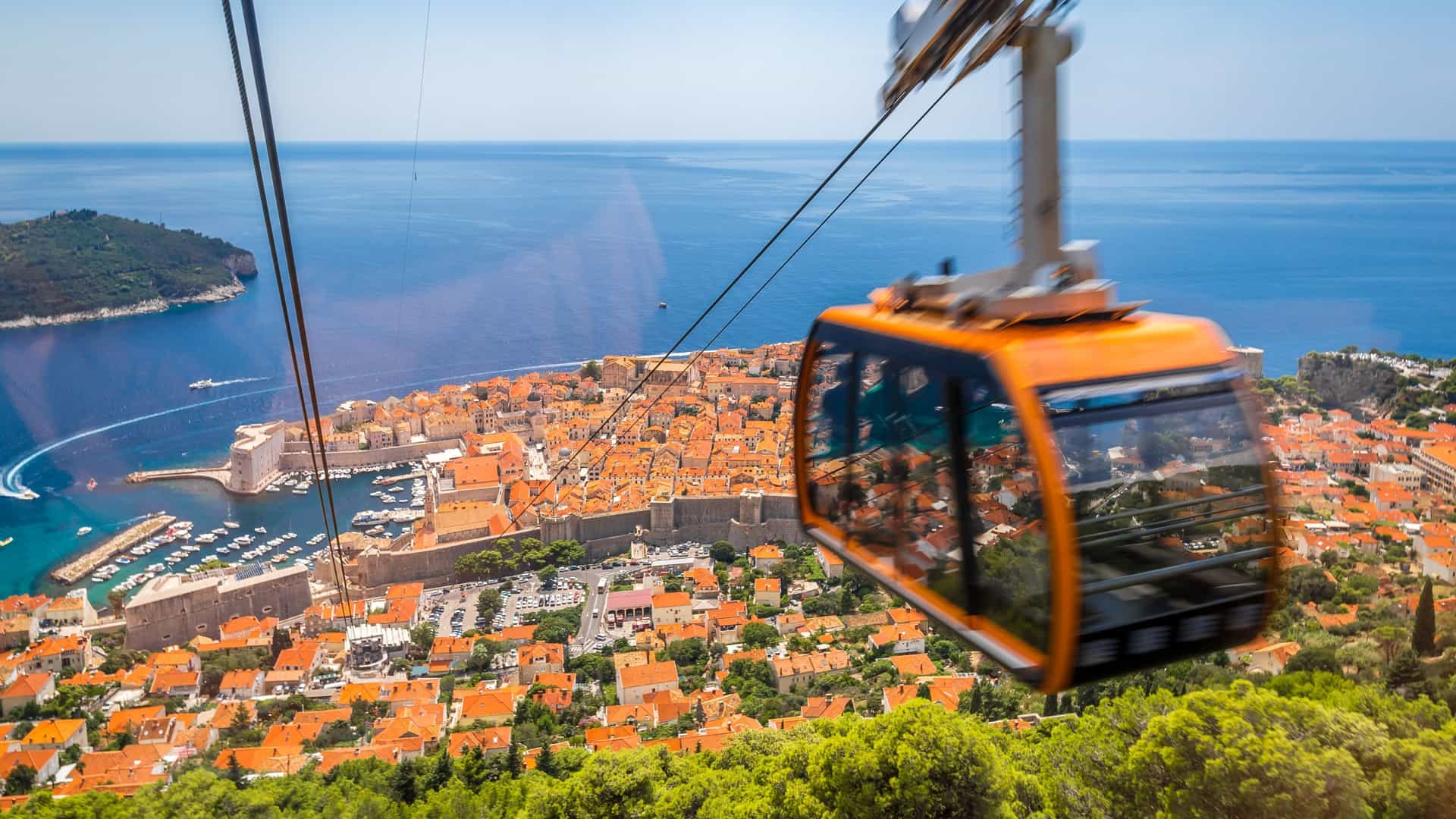 An orange Dubrovnik Cable Car car is seen traveling up a steep mountain with a lush green landscape. Below, the red tiled roofs and white stone walls of the historic Dubrovnik Old City are visible, surrounded by the deep blue Adriatic Sea and several small islands.