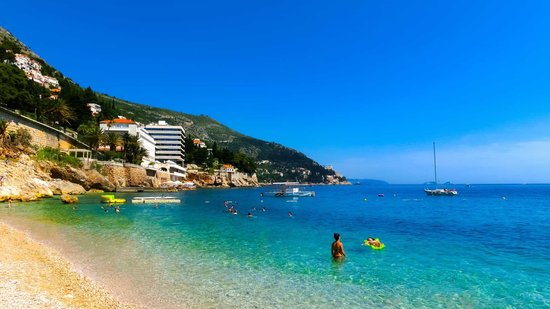 An aerial view of Banje Beach in Dubrovnik, showing a mixture of tourists relaxing on sunbeds and parasols on the pebble shore, while others are swimming and enjoying water activities in the crystal clear blue sea. The fortified walls of Dubrovnik's Old Town and the island of Lokrum are visible in the background.
