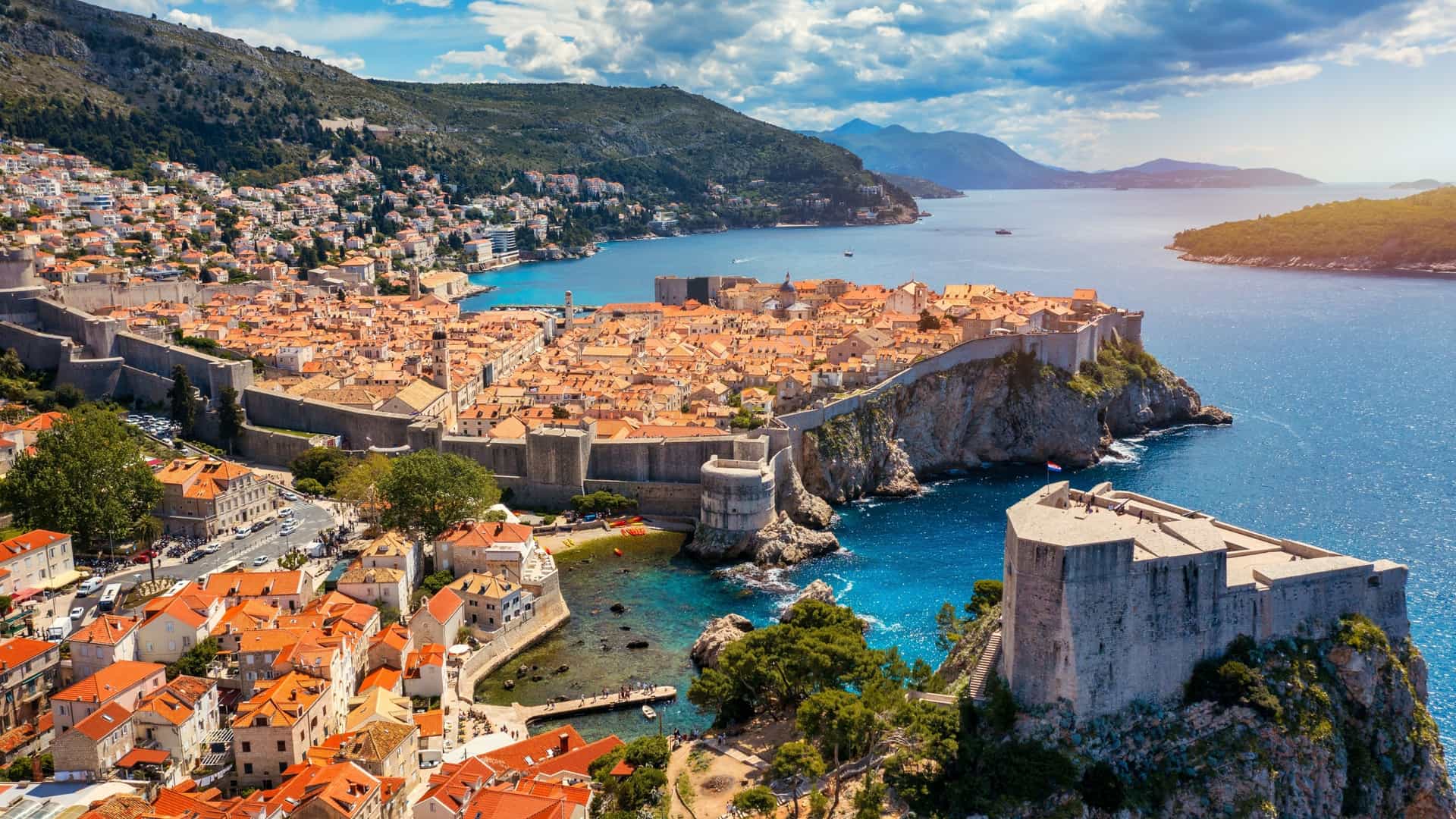 A view of Fort Lovrijenac, a historic fortress built on a high cliff overlooking the sea outside the city walls of Dubrovnik. The fortress has a triangular shape and is made of light colored stone, with a small archway visible near the base. The base of the cliff is surrounded by the blue sea.