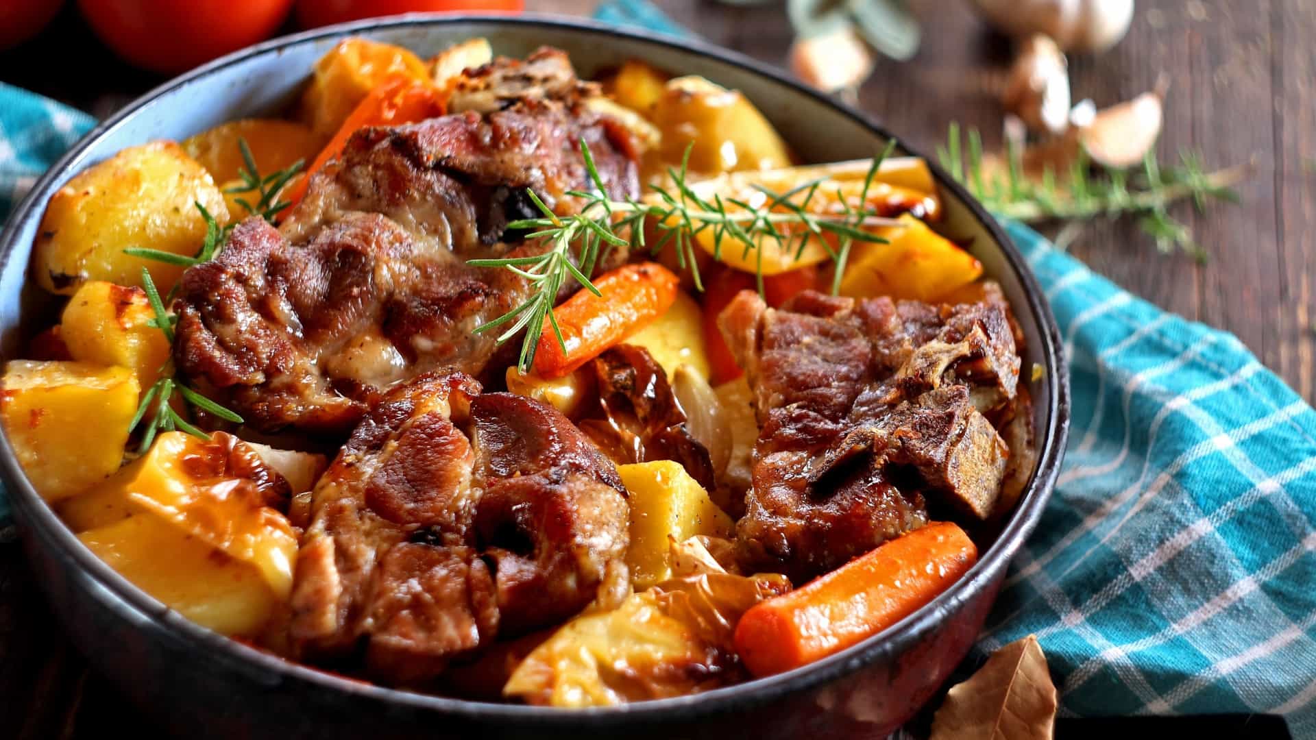 A close up view of a traditional Croatian peka dish, which is a slow cooked Pot Roast featuring large, tender pieces of meat (likely lamb or veal) nestled among roasted vegetables, including potatoes and carrots. The dish is served in a rustic bowl and garnished with fresh rosemary sprigs.