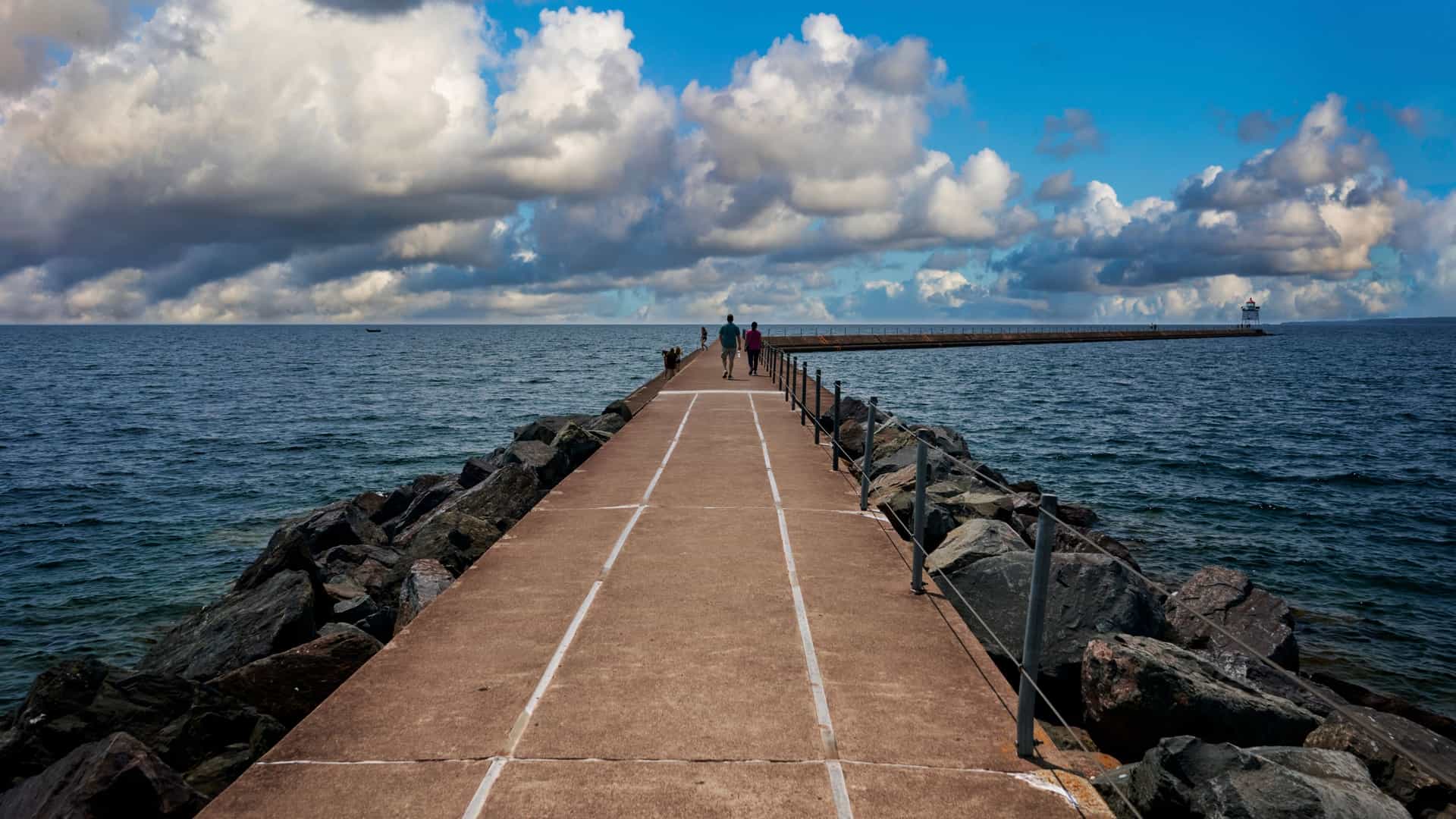 A wide angle, scenic view captures the Duluth, Minnesota, waterfront, where the city skyline meets the vast expanse of Lake Superior. The iconic Aerial Lift Bridge and harbor structures are visible, with the tranquil water reflecting the light under a bright, clear sky.