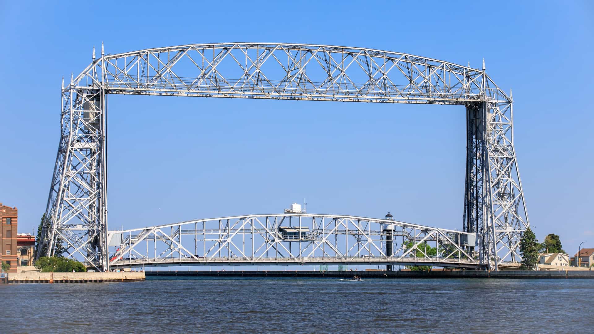The Aerial Lift Bridge in Duluth, Minnesota, is clearly visible spanning the canal entrance to the harbor. The massive steel structure is lowered, allowing a large freighter to pass directly underneath it as the ship makes its way out toward Lake Superior.