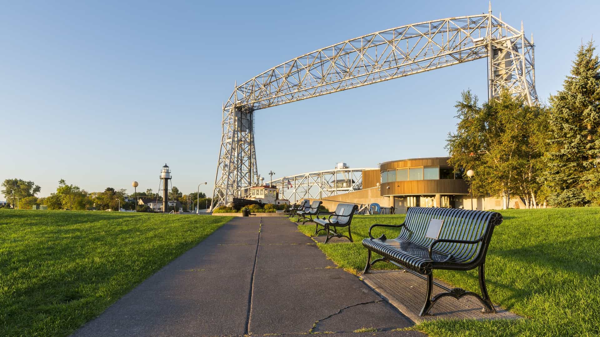 A paved sidewalk in Duluth's Canal Park is lined with numerous dark green park benches, offering visitors a place to rest. The perspective draws the eye straight down the path toward the massive steel structure of the Aerial Lift Bridge spanning the canal in the distance.