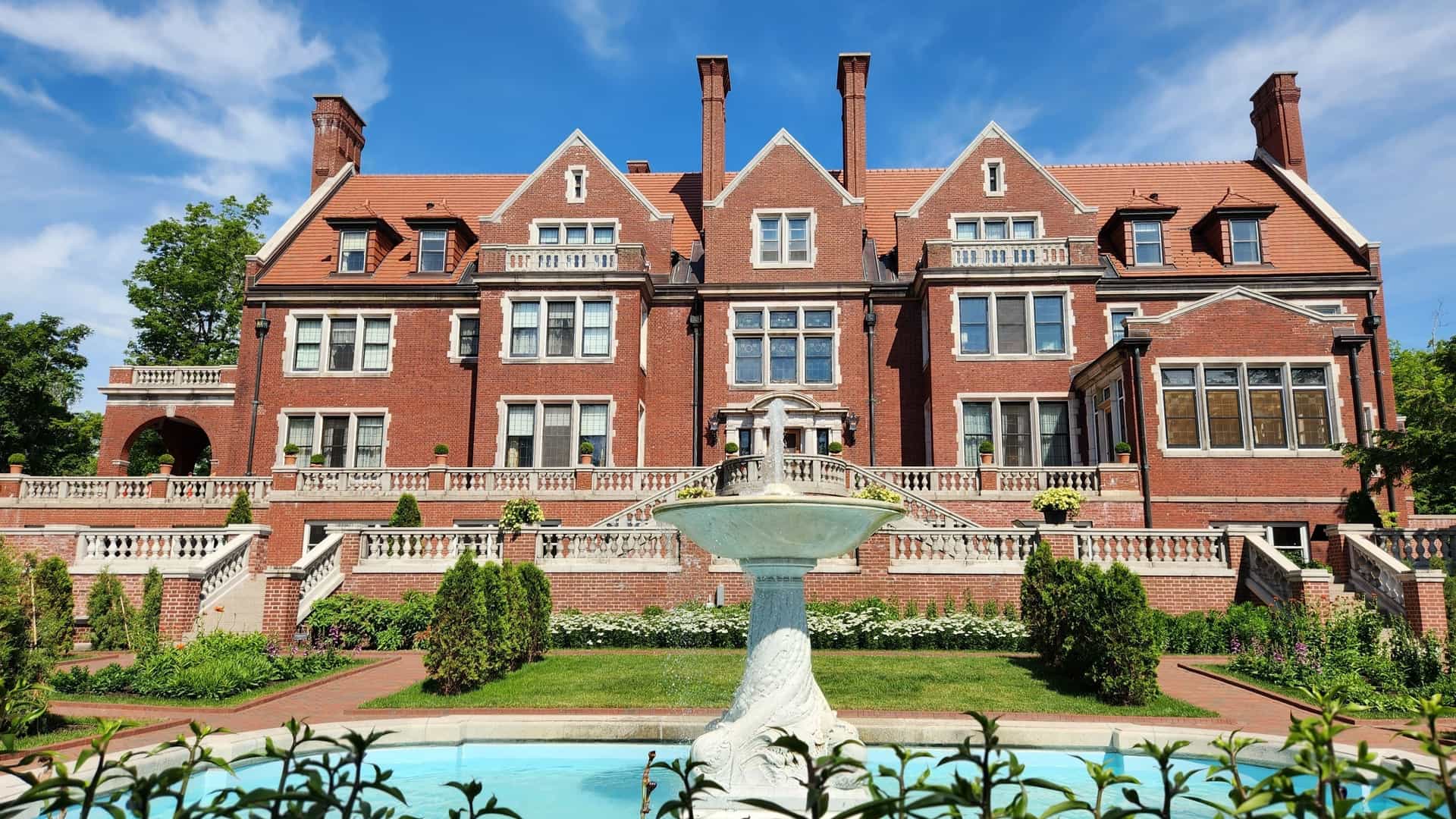 A large, elegant marble fountain featuring sculpted fish stands in the meticulously maintained backyard of the Glensheen Mansion in Duluth. Beyond the fountain, the grand, red brick Jacobean Revival manor house, with its numerous gables and chimneys, serves as a striking backdrop to the formal garden area.