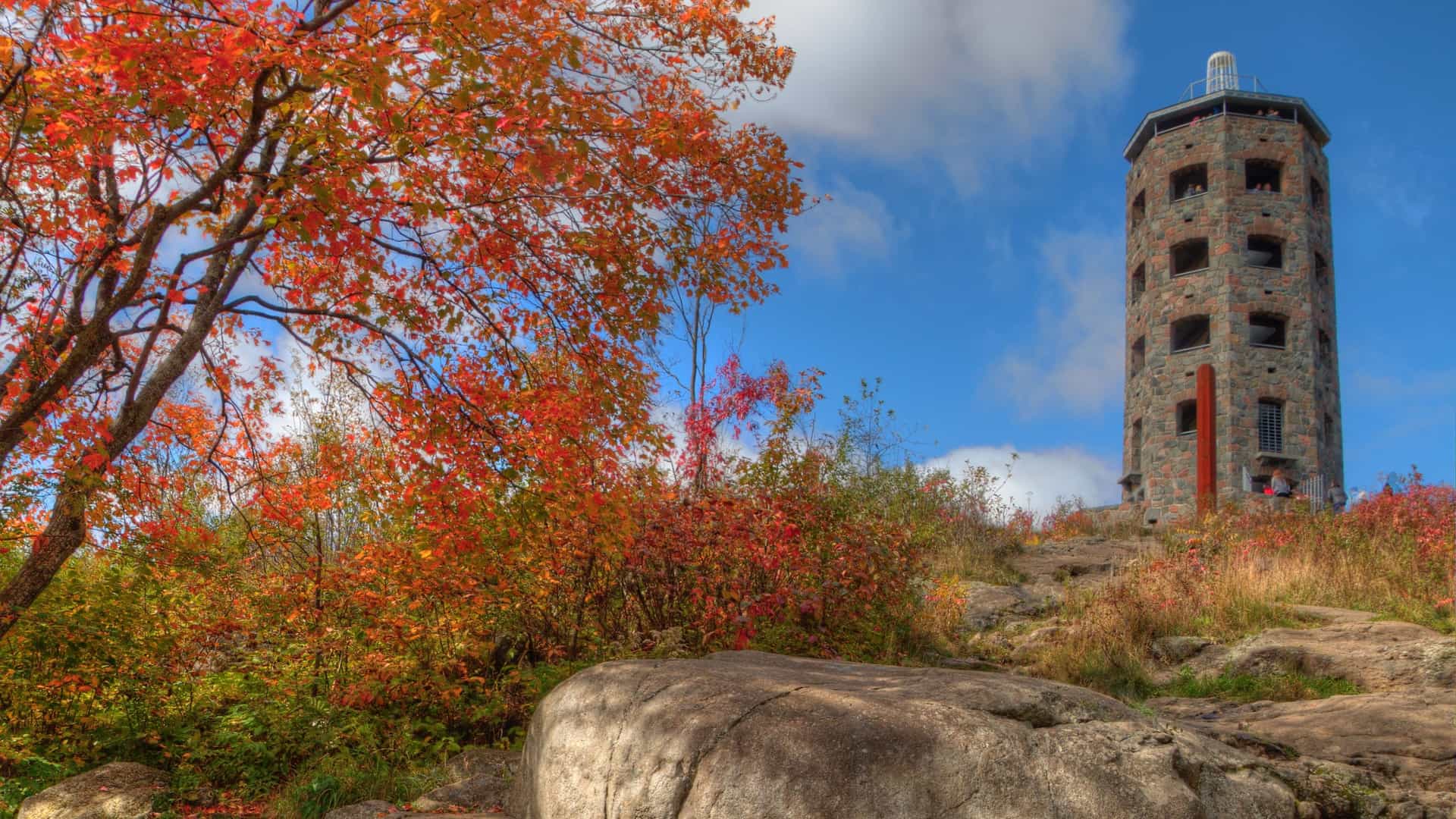 The five story Enger Tower, a gray stone observation tower, stands prominently atop Enger Hill in Duluth, Minnesota, serving as a popular tourist destination. From this high vantage point, the scenic view encompasses the city's harbor, the curve of Park Point, and the vast, blue expanse of Lake Superior.
