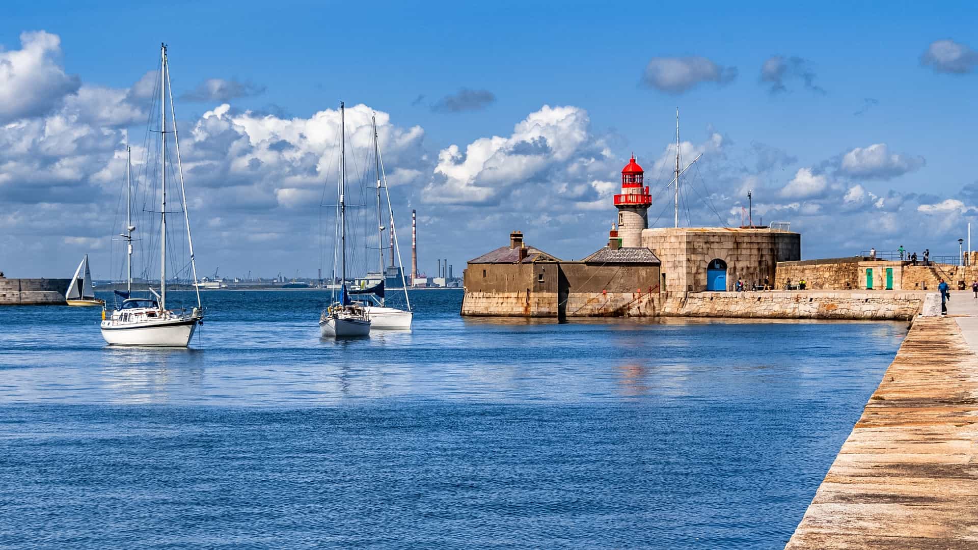 A detailed view along the granite surface of the Dun Laoghaire East Pier in Dublin, Ireland. The foreground is dominated by the wide, straight pier, which leads the eye toward the distinctive round, white lighthouse at its far end. The vast expanse of the harbor and the sea is visible to the side.