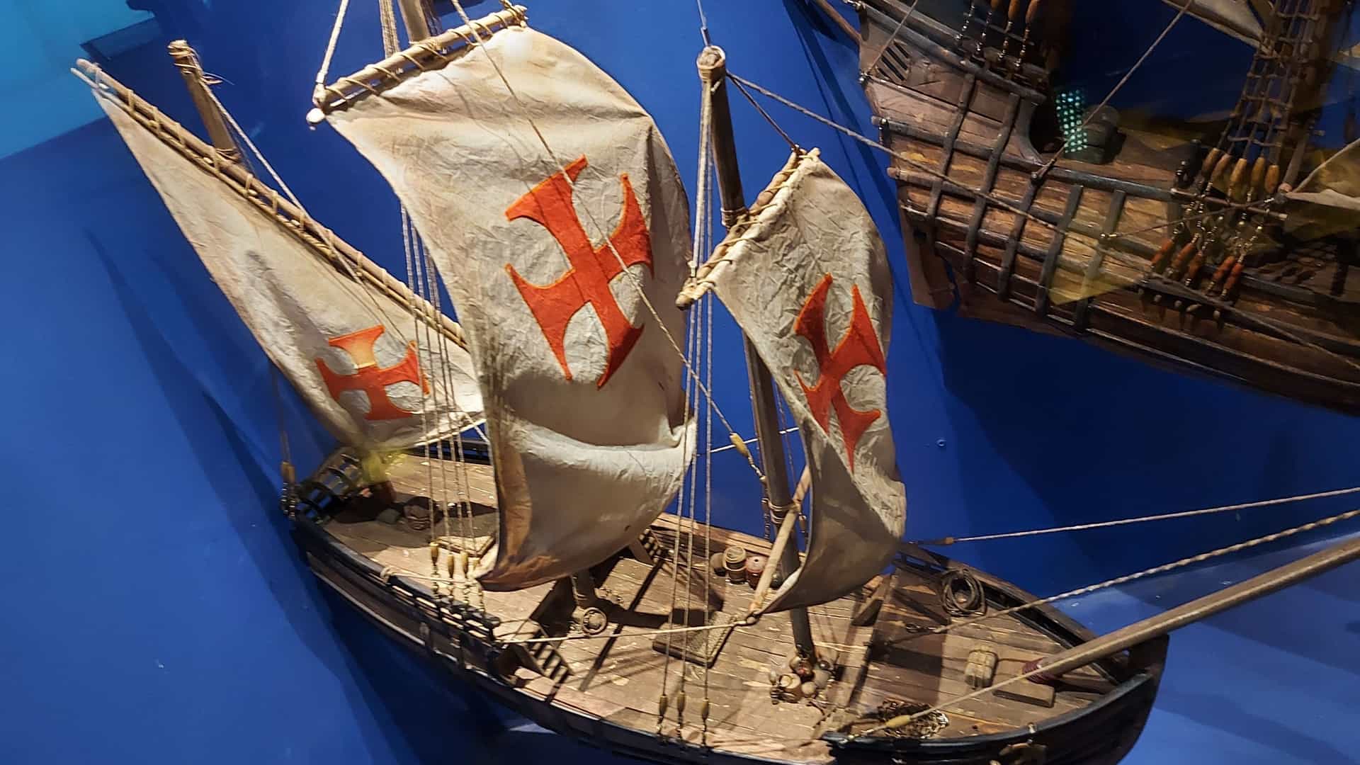 A detailed, high-angle view of a ship model displayed inside a museum, likely the National Maritime Museum of Ireland. The model depicts a historical caravel or similar vessel with billowing white sails marked prominently with a large, red Cross of the Order of Christ. The model rests on a deep blue background, simulating water, with the dark hull and wooden deck visible.