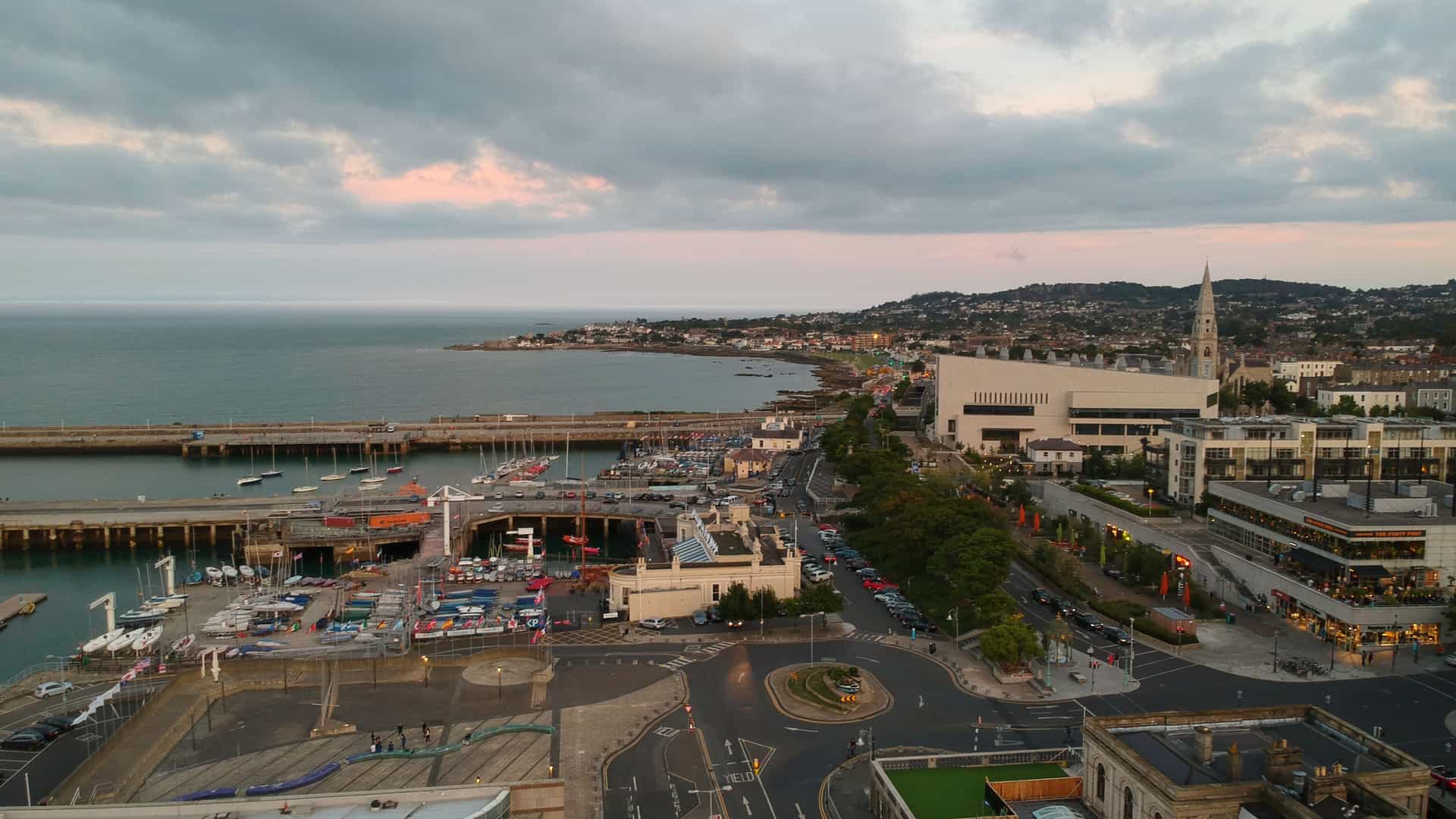An aerial view of the coastal town of Dún Laoghaire, Ireland, at dusk. The image features a busy harbor filled with boats and protected by a long stone pier, a central roundabout connecting the town's streets, and modern and historical buildings, all backed by a rolling hill.
