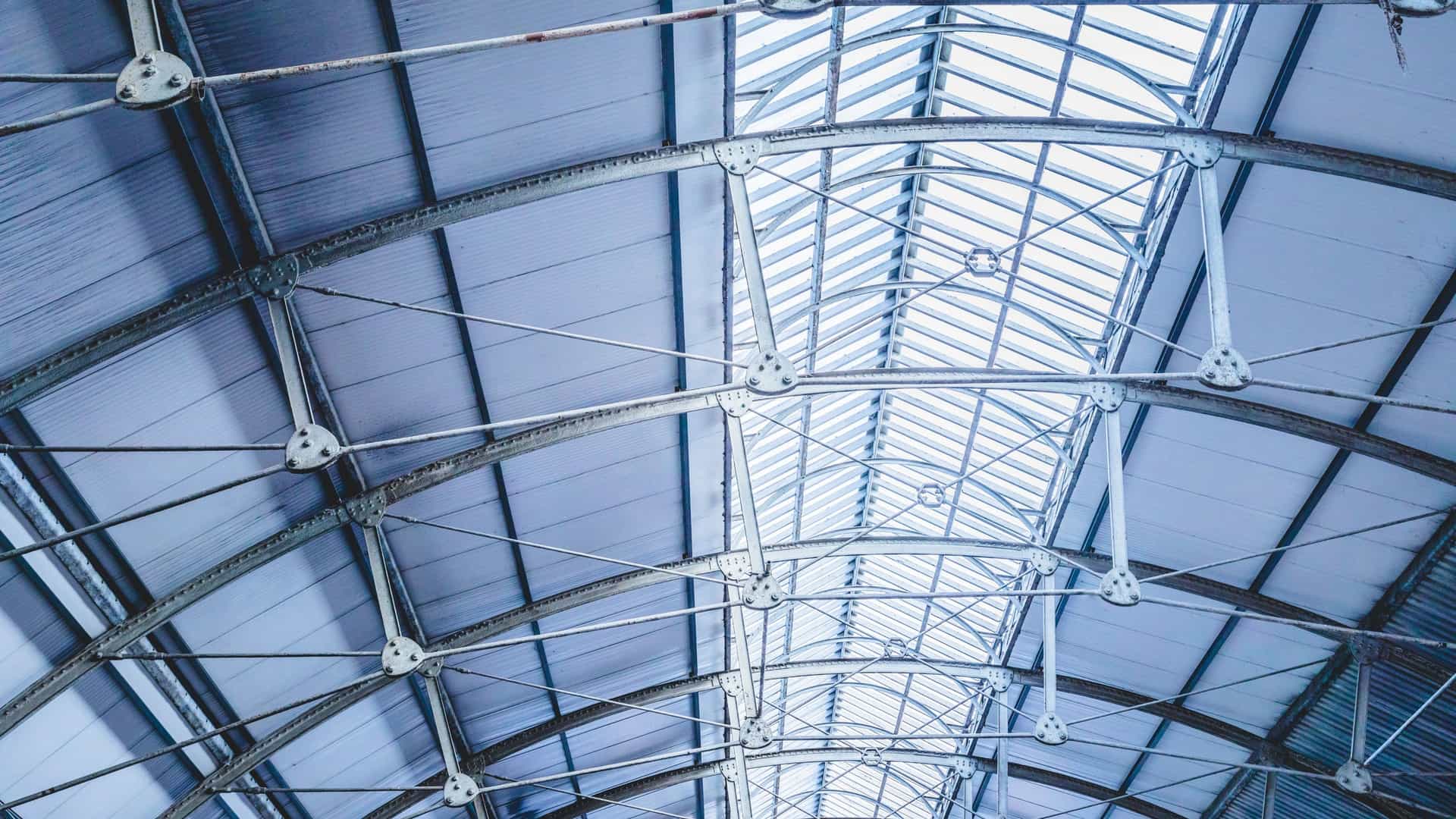 An upward-facing abstract view of the historic roof structure within a Dublin Area Rapid Transit (DART) station. The image highlights the detailed engineering of the vaulted ceiling, showing the curved, crisscrossing metal trusses with visible rivets and a central skylight running the length of the metal roof.