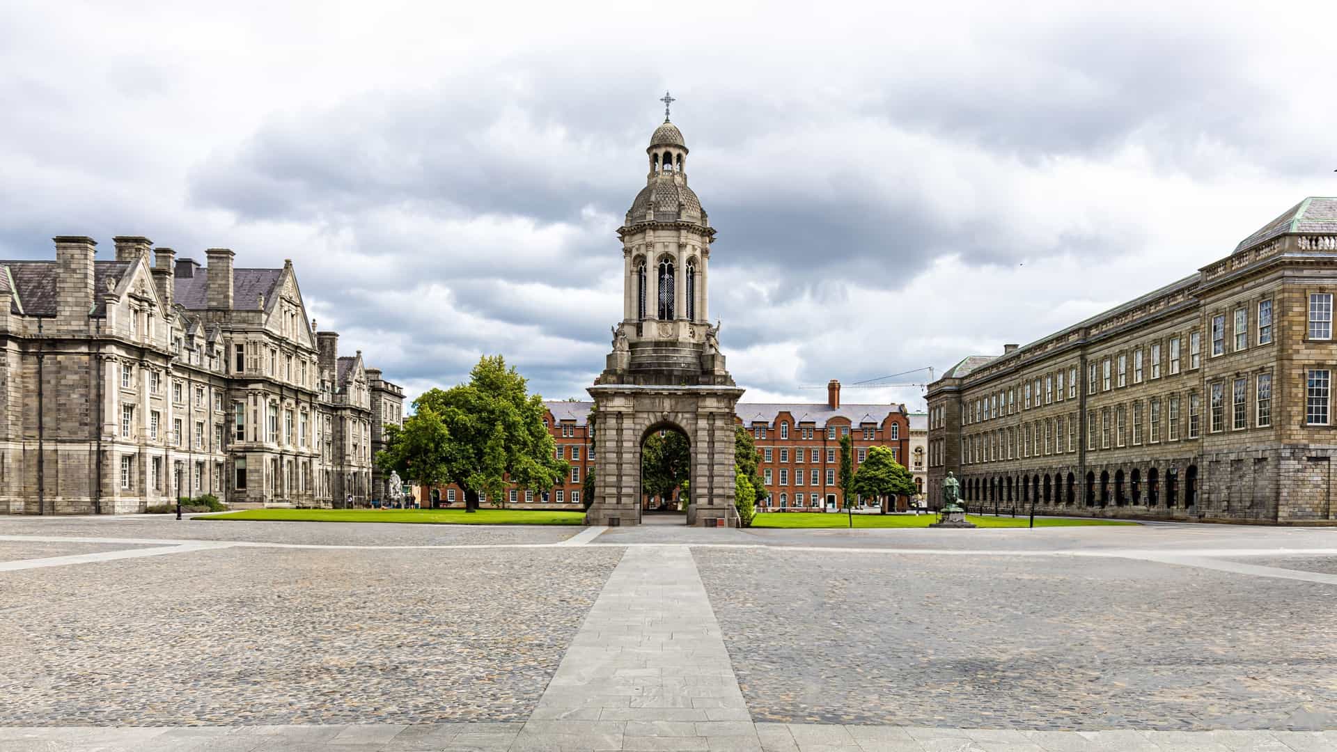 A panoramic view of the historic Trinity College Dublin campus, featuring the iconic Campanile bell tower standing tall in the main courtyard. Surrounded by lush green lawns and grand classical architecture, this image captures the beautiful and academic atmosphere of Ireland's oldest university on a bright, sunny day.