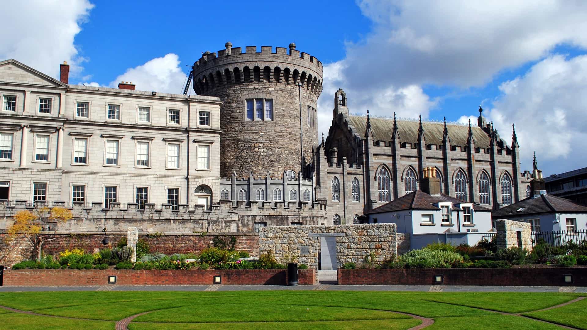 A sunlit, exterior view of the historic Dublin Castle in Ireland. The photograph prominently features the elegant Neobaroque architecture of the central complex, likely viewed from the large courtyard. This well-known Irish landmark is highlighted against a blue sky, emphasizing its importance as a center of governance and a prominent tourist attraction.
