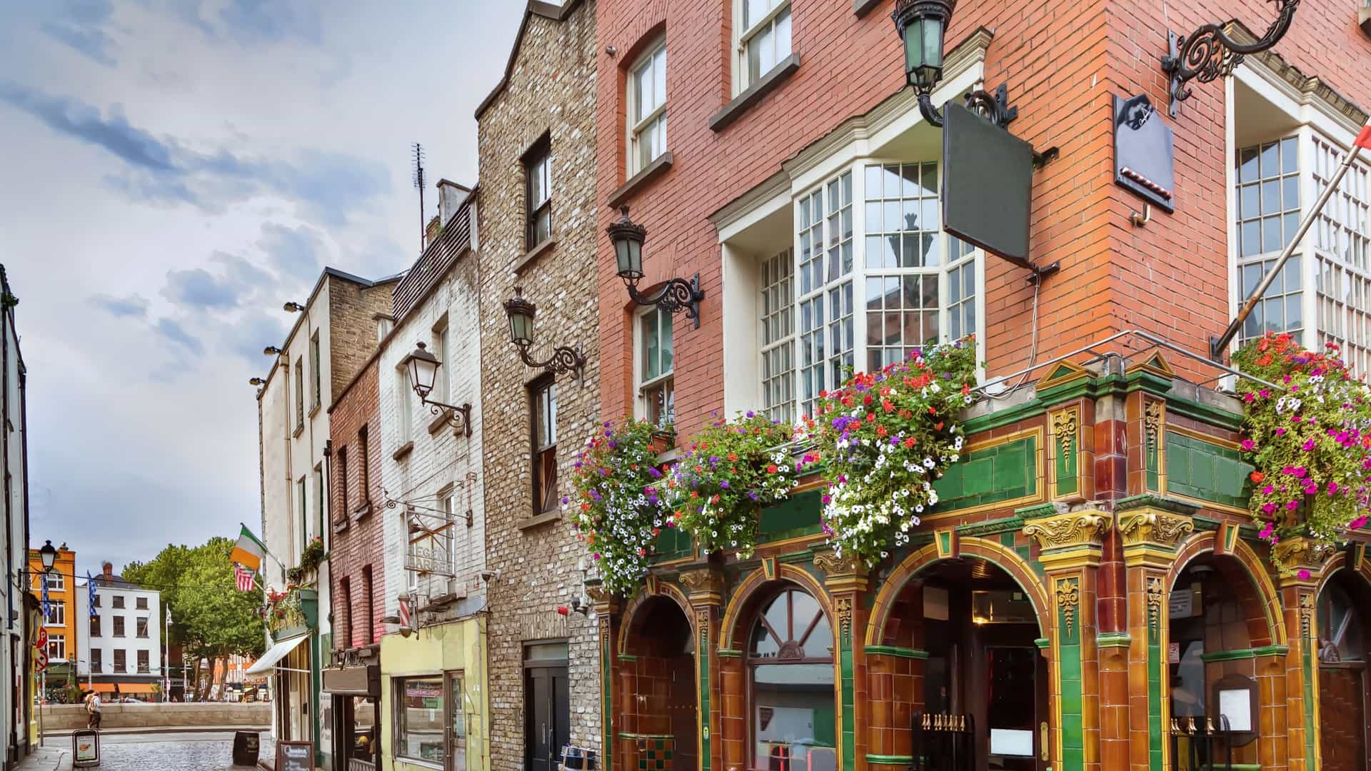 A photograph capturing the vibrant and historic Temple Bar street in Dublin City Centre, Ireland. The narrow, cobblestone thoroughfare is flanked by traditionally colorful Irish pubs and buildings. The scene is lively, likely bustling with tourists and locals enjoying the atmosphere of Dublin's famous cultural and nightlife quarter.
