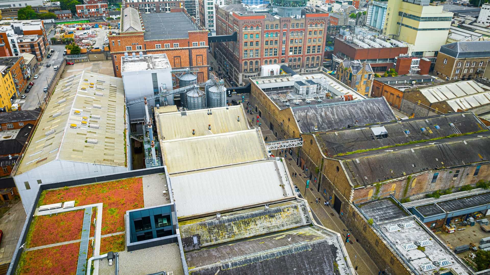 An aerial photograph showcasing the Guinness Storehouse, Ireland's most popular tourist attraction, within the sprawling St. James's Gate Brewery complex in Dublin. The image highlights the distinctive historic industrial buildings and the modern glass structure of the Storehouse, offering a view of the surrounding Dublin city landscape under a clear sky.