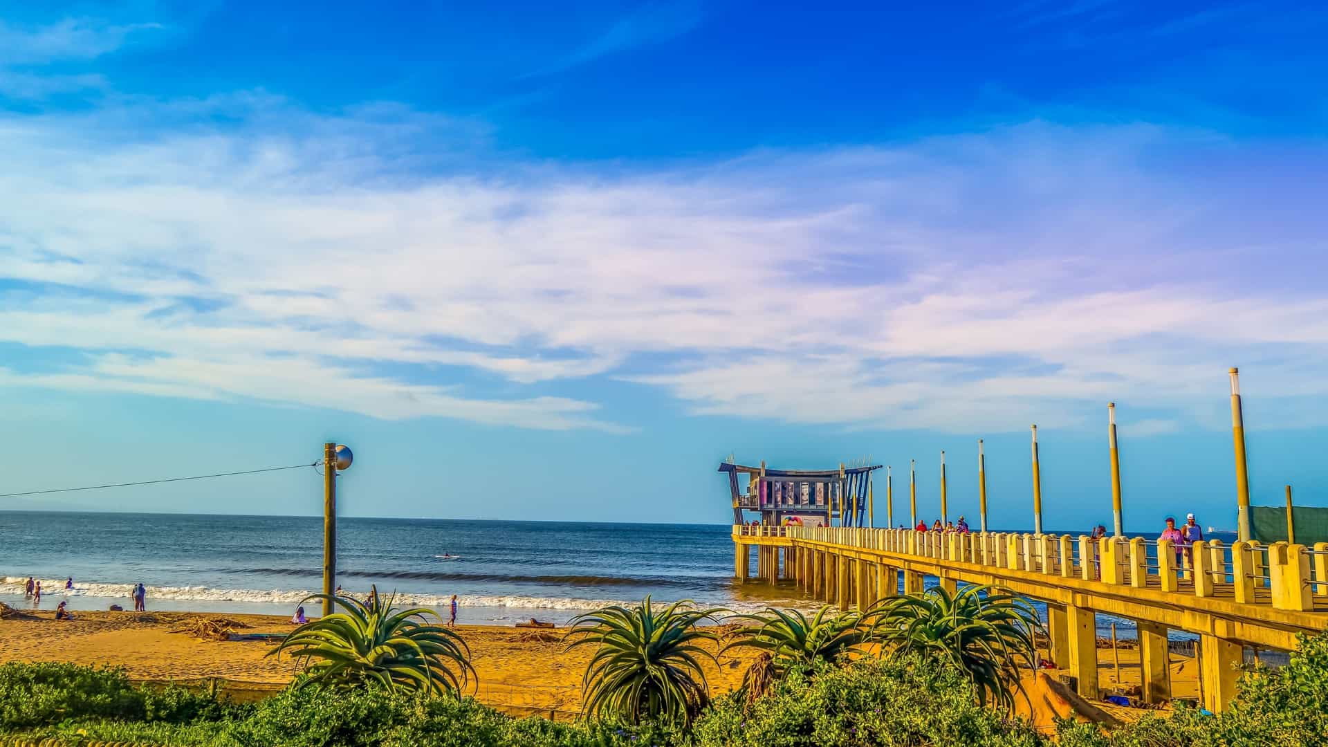 The curving pier extending into the Indian Ocean along Durban's Golden Mile, specifically at uShaka Beach in Durban, South Africa, with the vast sandy beach and cityscape visible in the background.