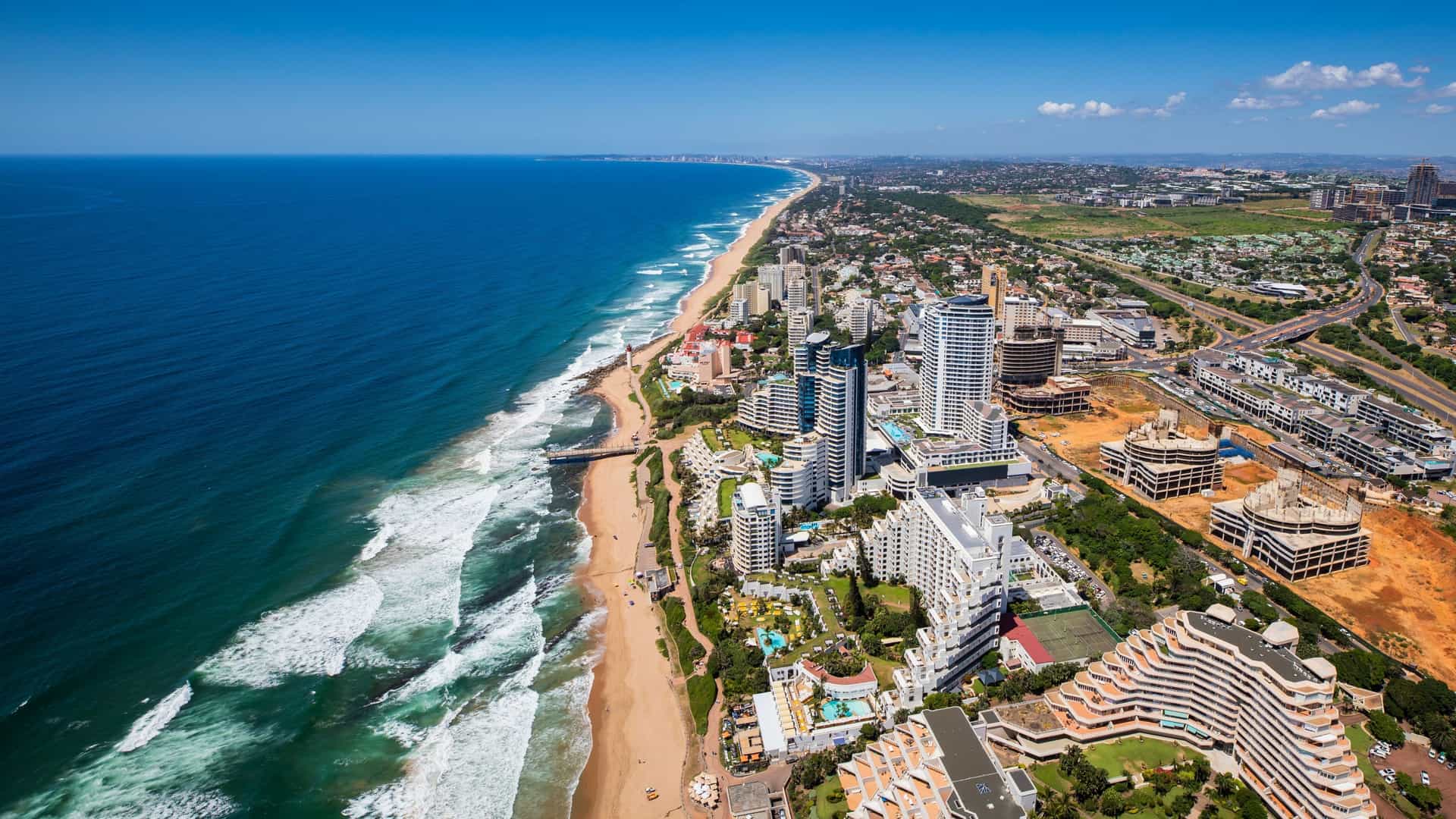 The iconic Umhlanga Rocks Lighthouse stands on the coastline of Umhlanga Rocks, near Durban, South Africa. The image features the red and white tower set against a clear sky and the ocean.