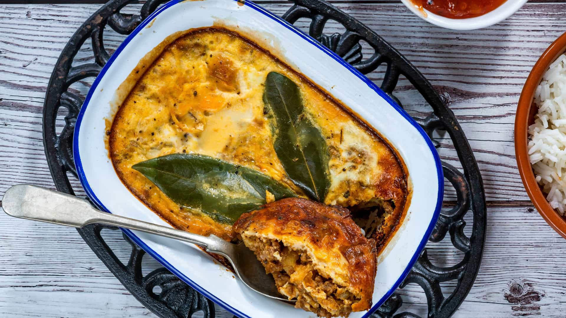 A dish of Bobotie, the traditional South African dish of curried minced meat baked with an egg-and-milk custard topping, served with yellow rice and dried fruit, possibly raisins or apricots.