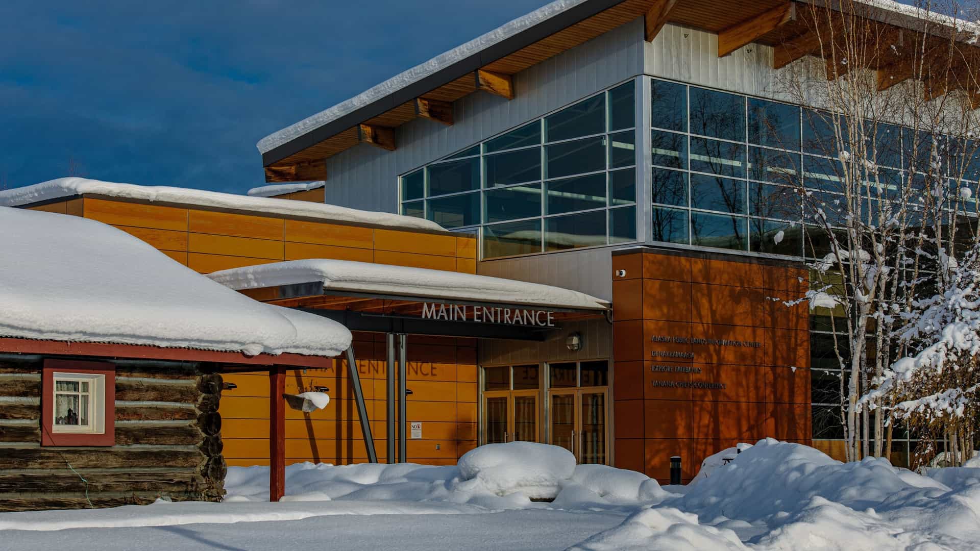 A striking wide-angle photograph capturing the Morris Thompson Cultural and Visitors Center in Fairbanks, Alaska, a modern, multi-story structure featuring a blend of natural wood, stone, and large glass windows. The architectural style incorporates Indigenous Alaska Native design elements. The image also prominently features the building's signature outdoor installation, the Antler Arch, created from hundreds of moose and caribou antlers, standing on the landscaped grounds in front of the building.