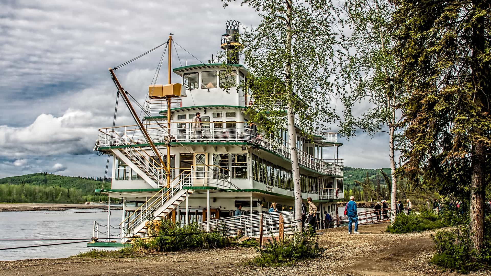 A three-story white sternwheeler paddleboat, identified as the Riverboat Discovery III, is docked on a river in Fairbanks, Alaska, likely the Chena River. The boat is a prominent tourist vessel with multiple decks, large windows, and an authentic paddlewheel visible at the stern. The scene likely includes the riverbank and surrounding trees under a blue sky.