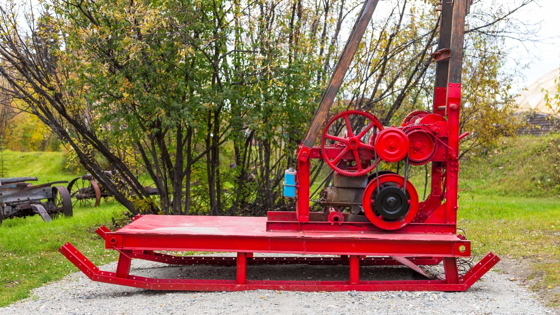 A collection of historic gold mining equipment is displayed outdoors in Pioneers Park (formerly Alaskaland) in Fairbanks, Alaska. The foreground features a pile of large, scoop-shaped, heavy-duty steel dredge buckets—the "shovels" of a gold dredge—next to a smaller, metal hand cart or mine car. The equipment is rusty and weathered, standing on gravel ground, and serves as a monument to the region's rich gold rush and mining history.