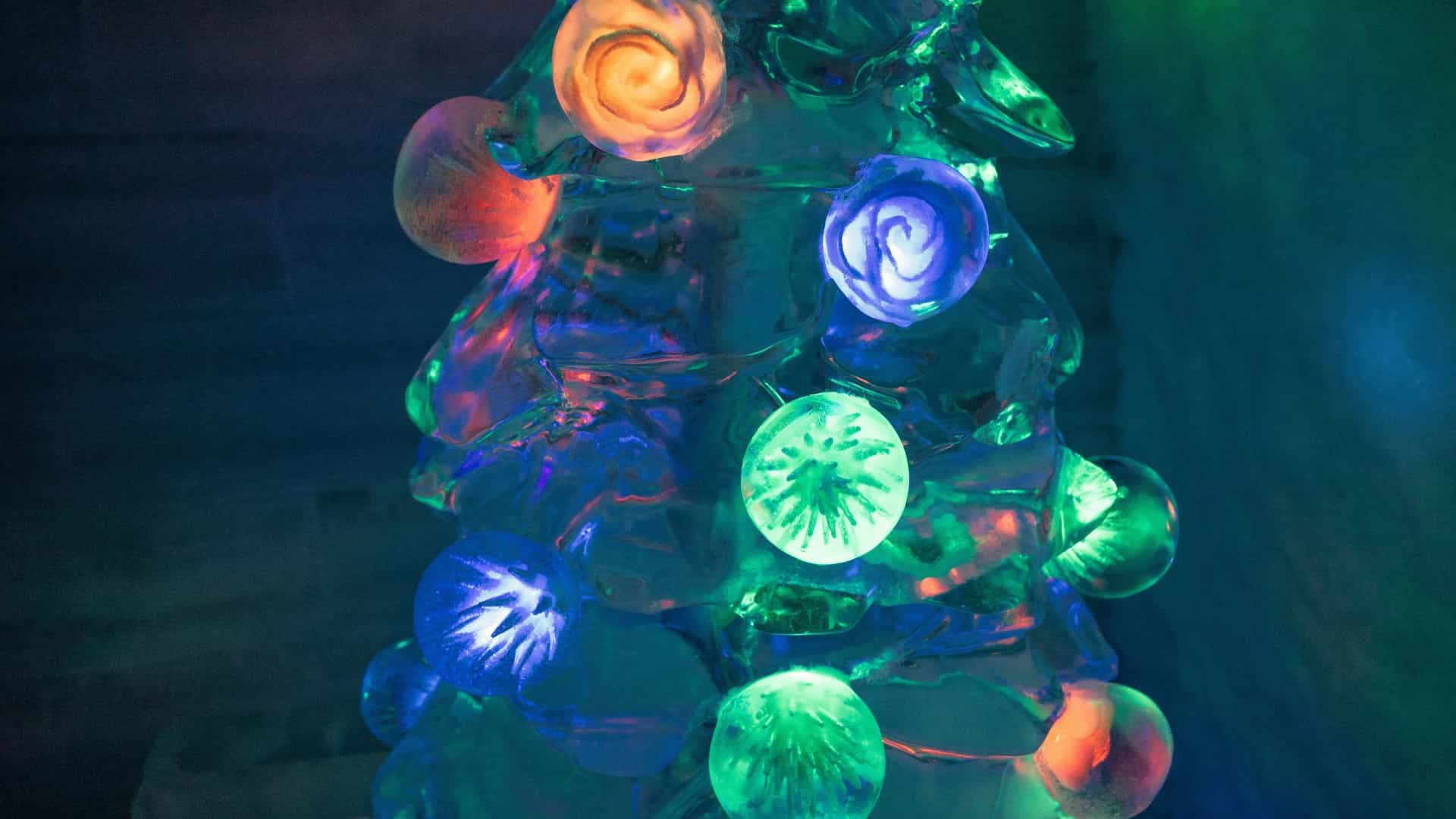 A vibrant, wide-angle interior view of the Aurora Ice Museum at Chena Hot Springs Resort near Fairbanks, Alaska. The image showcases the large, central ice bar, which is carved entirely from ice and illuminated by colored lights, primarily green and blue, giving the space a cool, ethereal glow. Ice sculptures and columns are visible throughout the foreground, and the overall environment is composed of thick, permanent ice. The date mentioned in the image's source (Mar 20, 2023) is a common time for winter tourism in Fairbanks.