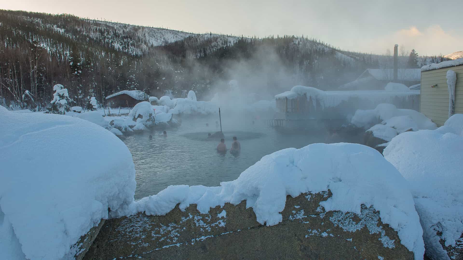 A sweeping, elevated view of the Chena Hot Springs Resort complex in Alaska's Interior, likely captured during the winter. The image shows the collection of resort buildings (lodges, cabins, and facilities) situated in a valley, with the surrounding snow-covered mountains and Alaskan wilderness prominent in the background. Steam from the geothermal operations and the hot springs pool often rises into the cold air, creating a distinctive atmosphere across the property.