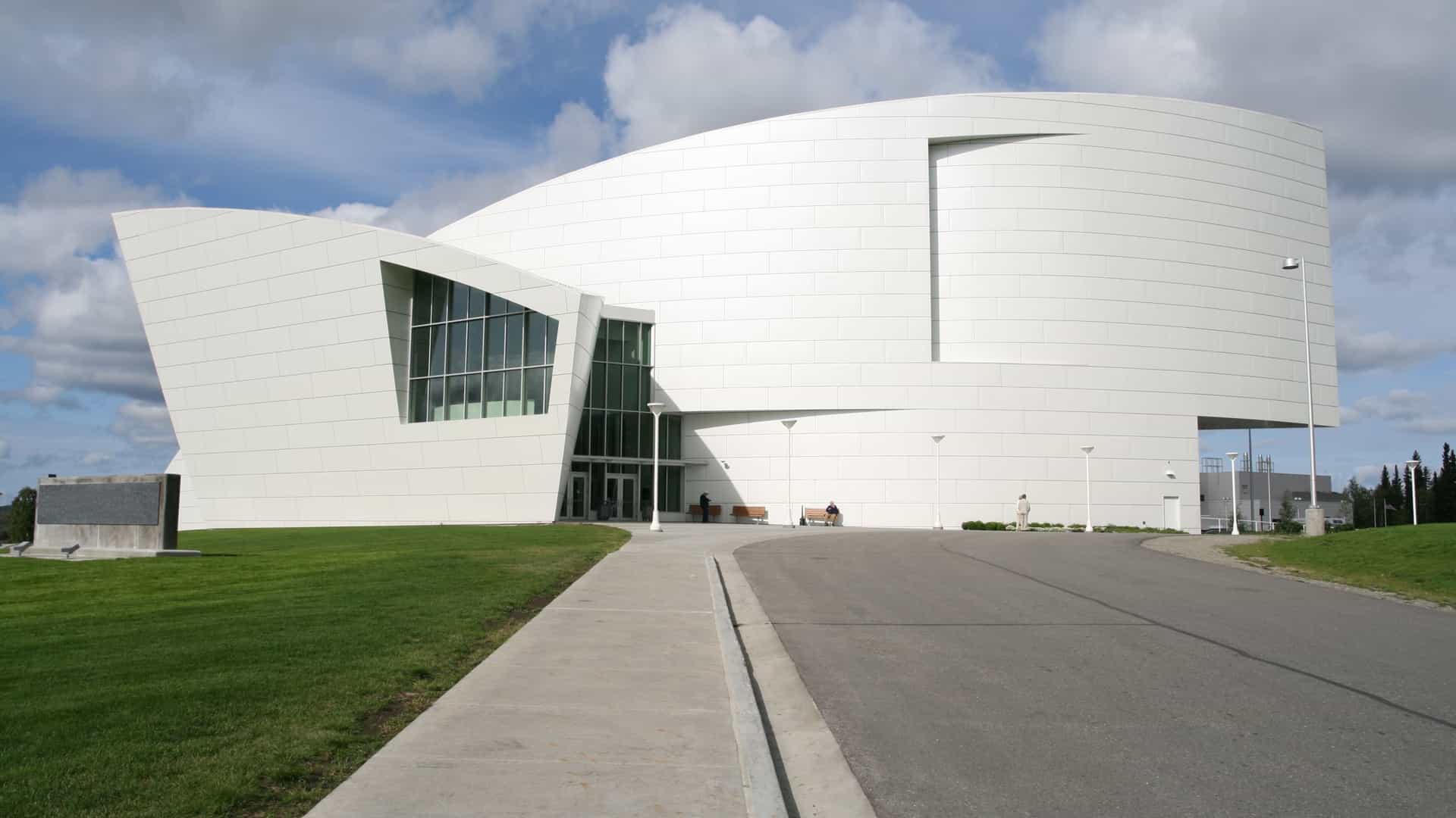 An exterior view of the modern, distinctive building that houses the University of Alaska Museum of the North in Fairbanks, Alaska. The unique, sweeping architecture is set against a backdrop of trees and a bright sky.