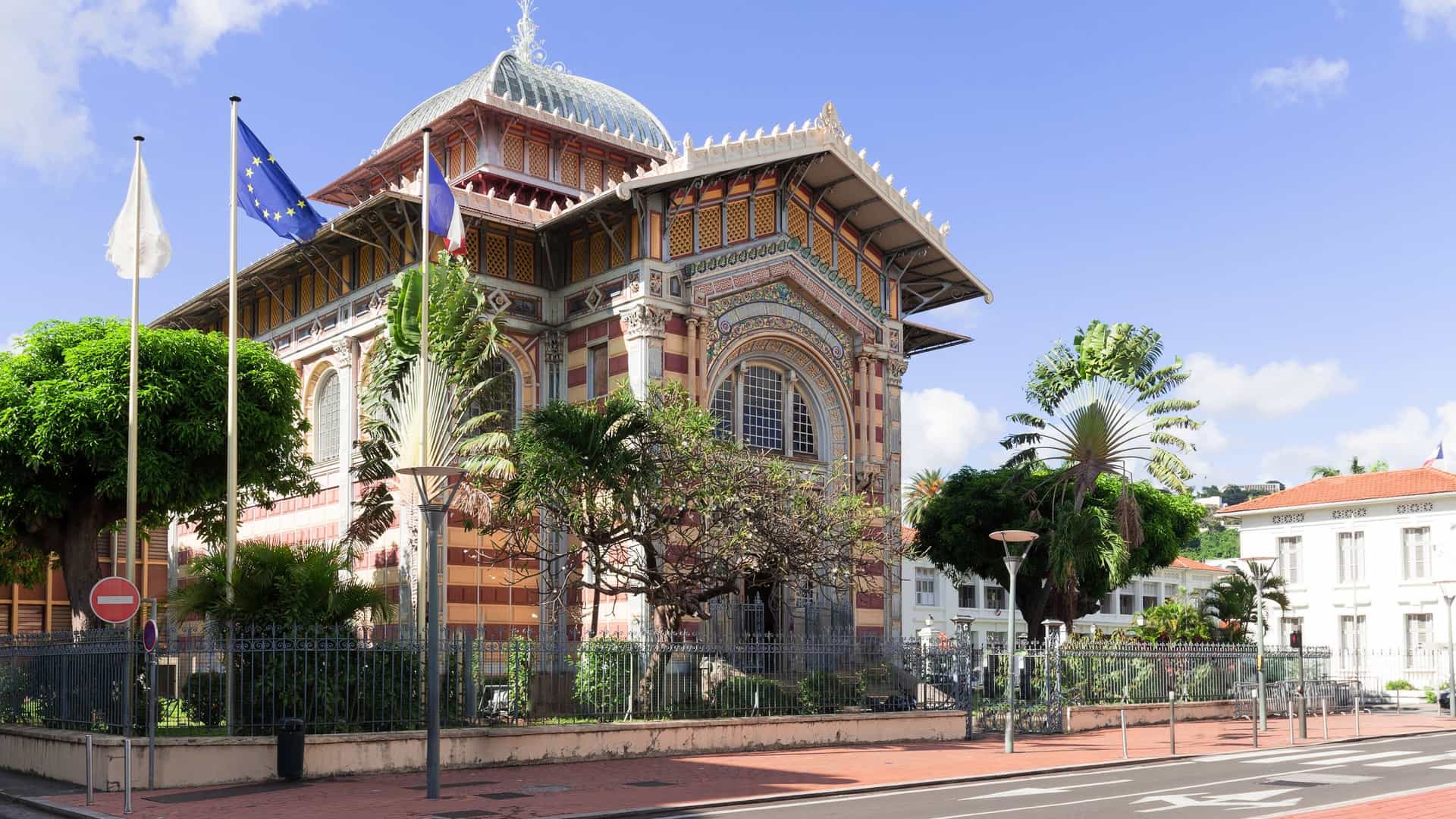 A wide-angle exterior view of the historic Schoelcher Library in Fort-de-France, Martinique. The intricate, glass-and-steel structure was originally built in Paris for the 1889 World's Fair before being disassembled and re-erected on the Caribbean island.