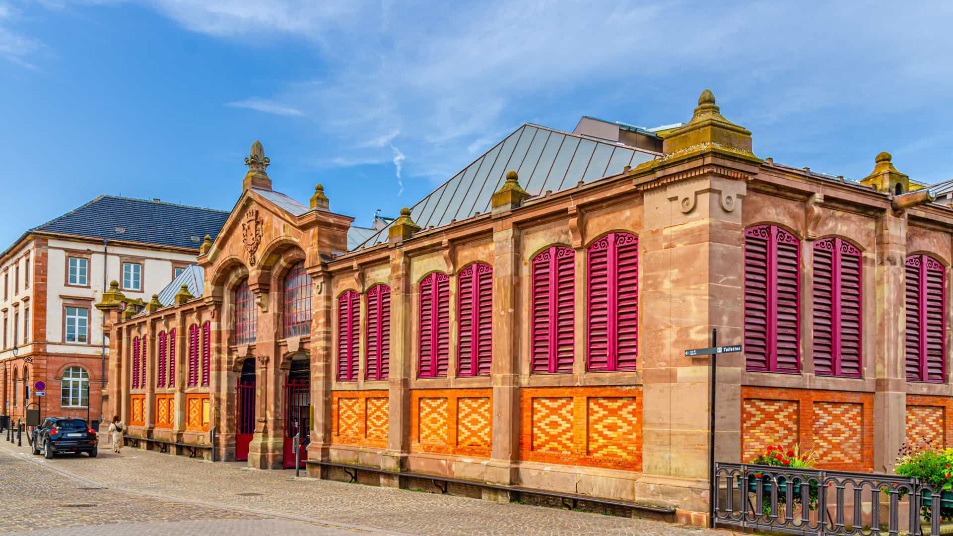 The image is a striking exterior view of the Covered Market (Marché Couvert) in Fort-de-France, Martinique. The building is a notable example of French Neo-Baroque architecture, featuring an ornate facade, elaborate ironwork, and a distinctive domed structure.