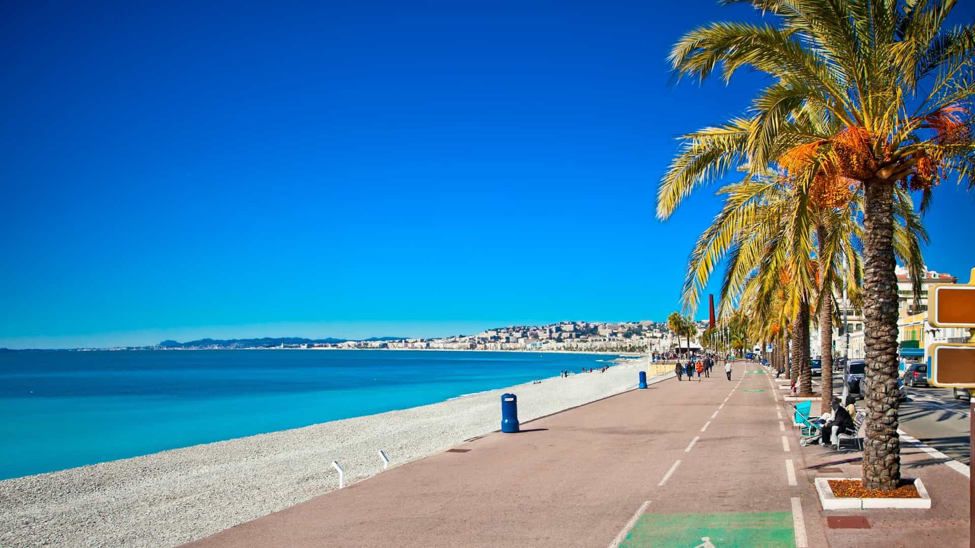 A scenic view of the Promenade des Anglais in Nice, France, a popular seafront promenade lined with palm trees. The blue Mediterranean Sea is on one side, and the city buildings are on the other.