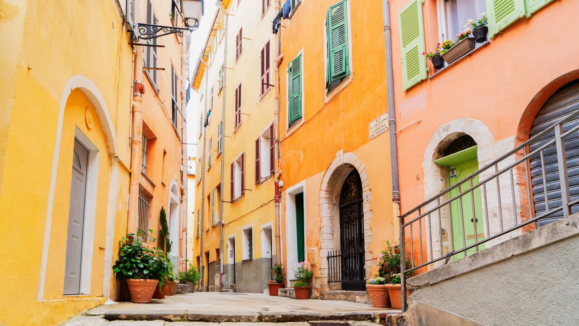 A vertical, street-level view of a narrow, cobbled alleyway in the Old Town (Vieux Nice), France. The street is lined with tall, historic apartment buildings featuring pastel-colored stucco facades in shades of ochre, terracotta, and yellow. Green wooden shutters frame the windows and laundry lines with clothes are strung between the balconies overhead, giving the scene a charming, Mediterranean atmosphere.