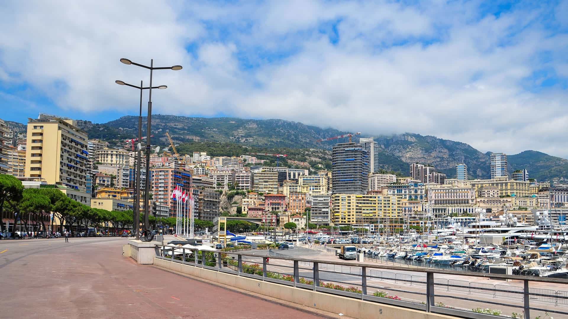 A sweeping, high-angle panoramic view of the city-state of Monaco, specifically focusing on the dense urban area of Monte Carlo and Port Hercule. The foreground shows a mass of high-rise apartments and hotels climbing the steep hillside, with the Prince's Palace on The Rock (Monaco-Ville) visible on the far left. The Port Hercule harbor is filled with rows of luxury yachts and boats moored in the bright turquoise Mediterranean Sea. The cityscape is set against the dramatic backdrop of the Maritime Alps rising in the distance under a clear blue sky.